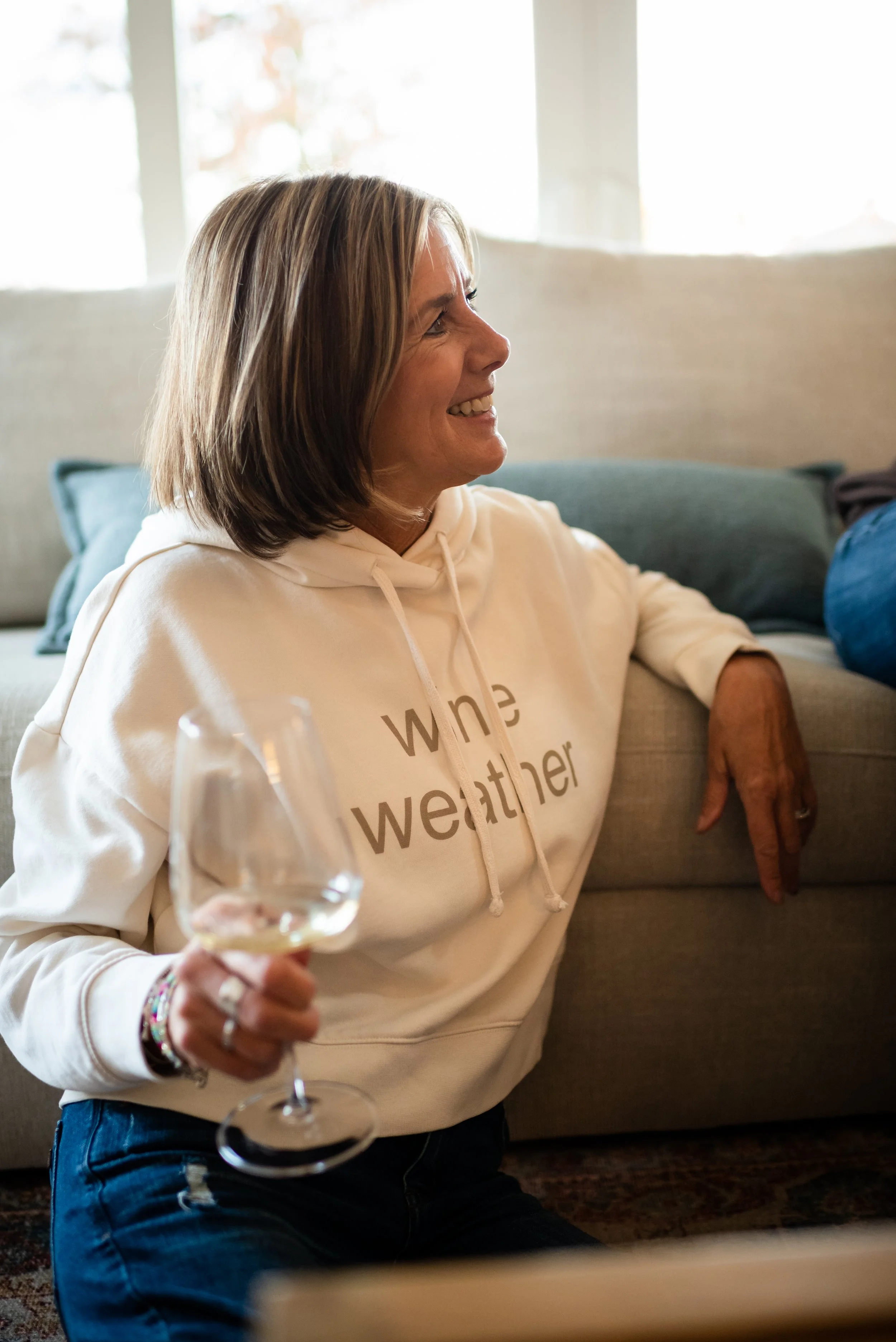 A woman with shoulder-length hair smiling while sitting on the floor, holding a glass of white wine in her right hand, wearing a cream hoodie that says 'wine weather,' in a cozy living room with a couch and cushions behind her.