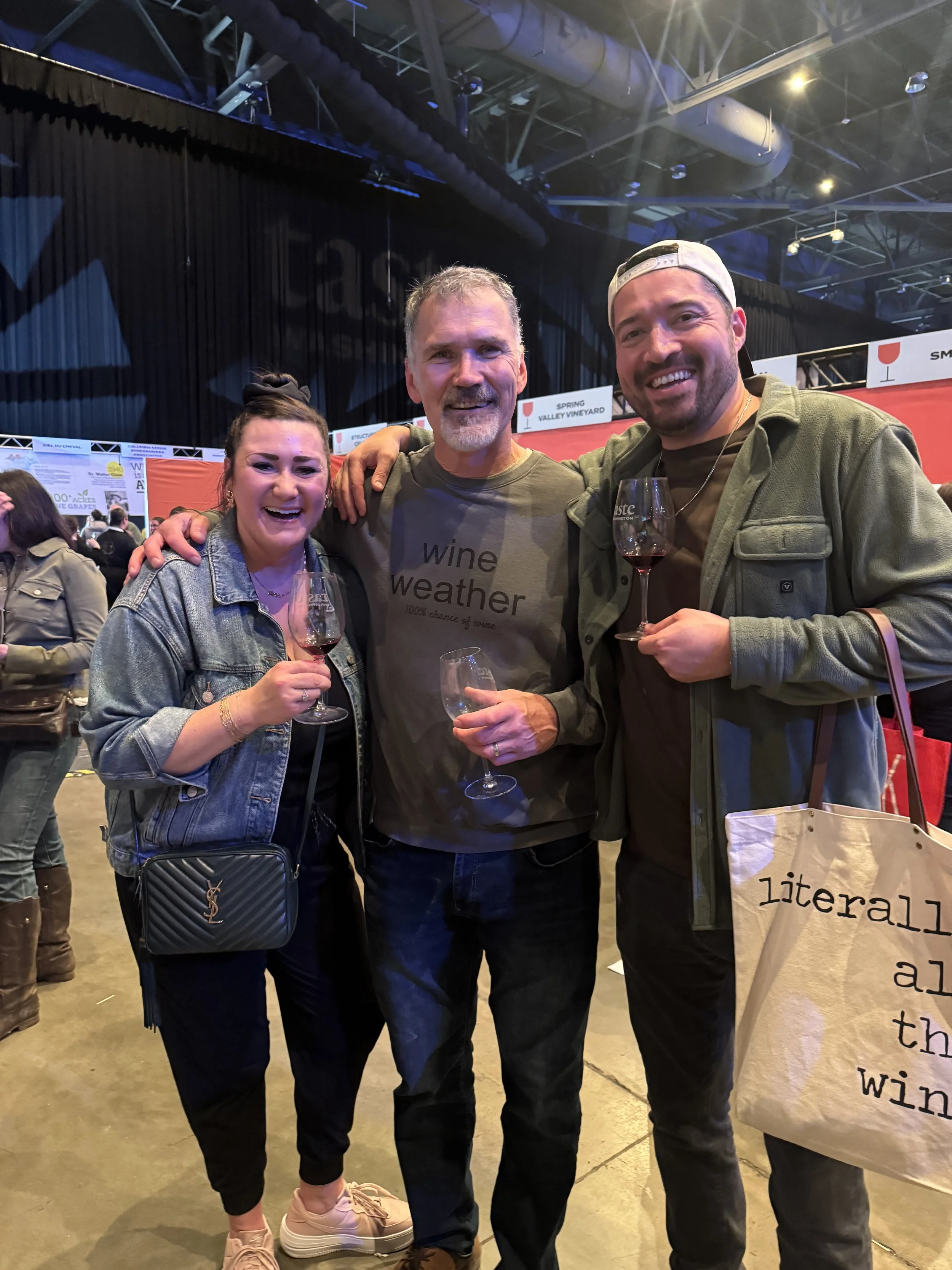 Three people smiling and holding wine glasses at an indoor event, with a woman on the left wearing a denim jacket and a purse, a man in the middle wearing a t-shirt that says 'wine weather,' and a man on the right wearing a cap and carrying a tote bag that says 'literally all the wine.'