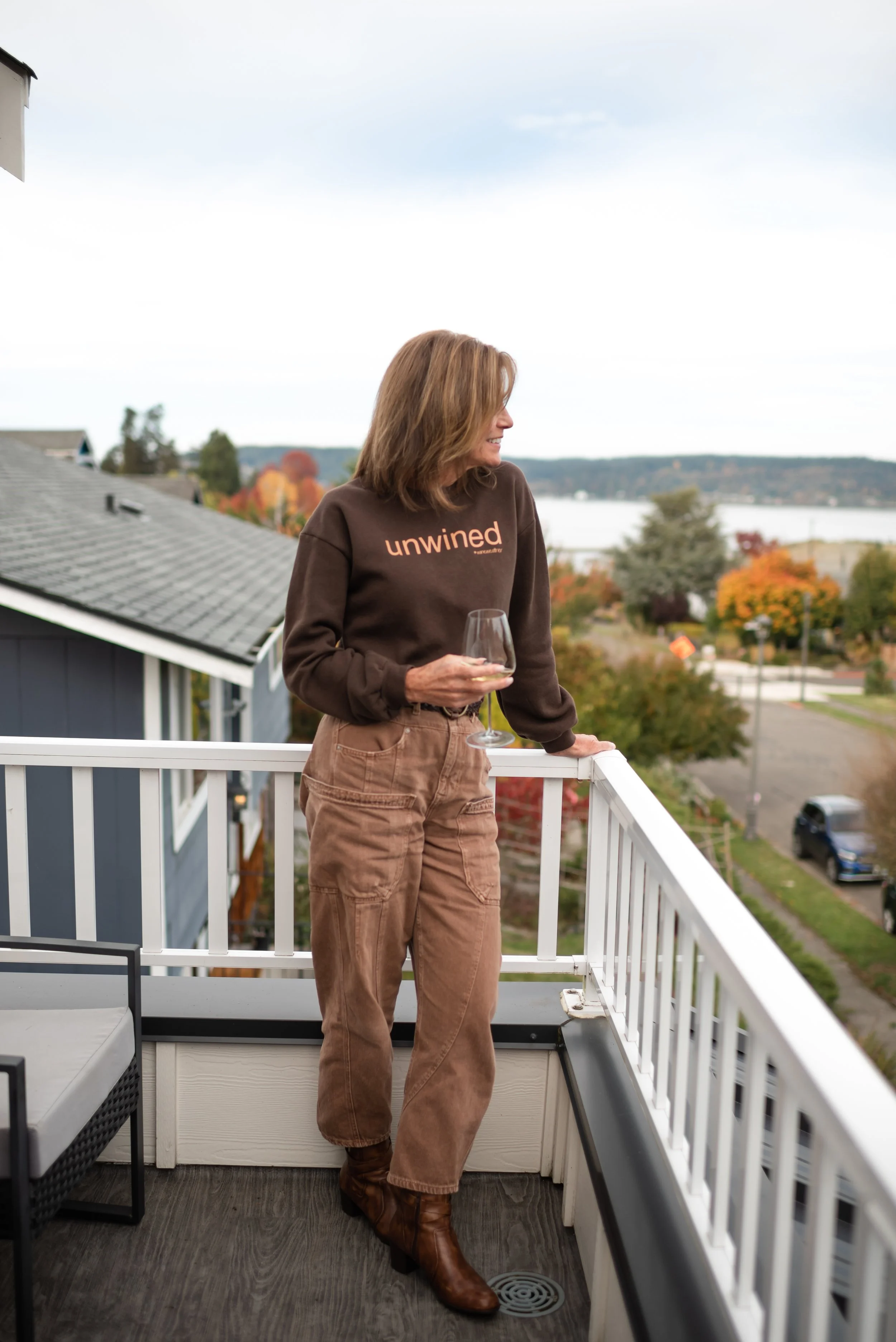 A woman standing on a balcony holding a glass of white wine, overlooking autumn trees and a body of water, wearing a brown sweatshirt with 'unwined' printed on it and brown cargo pants.