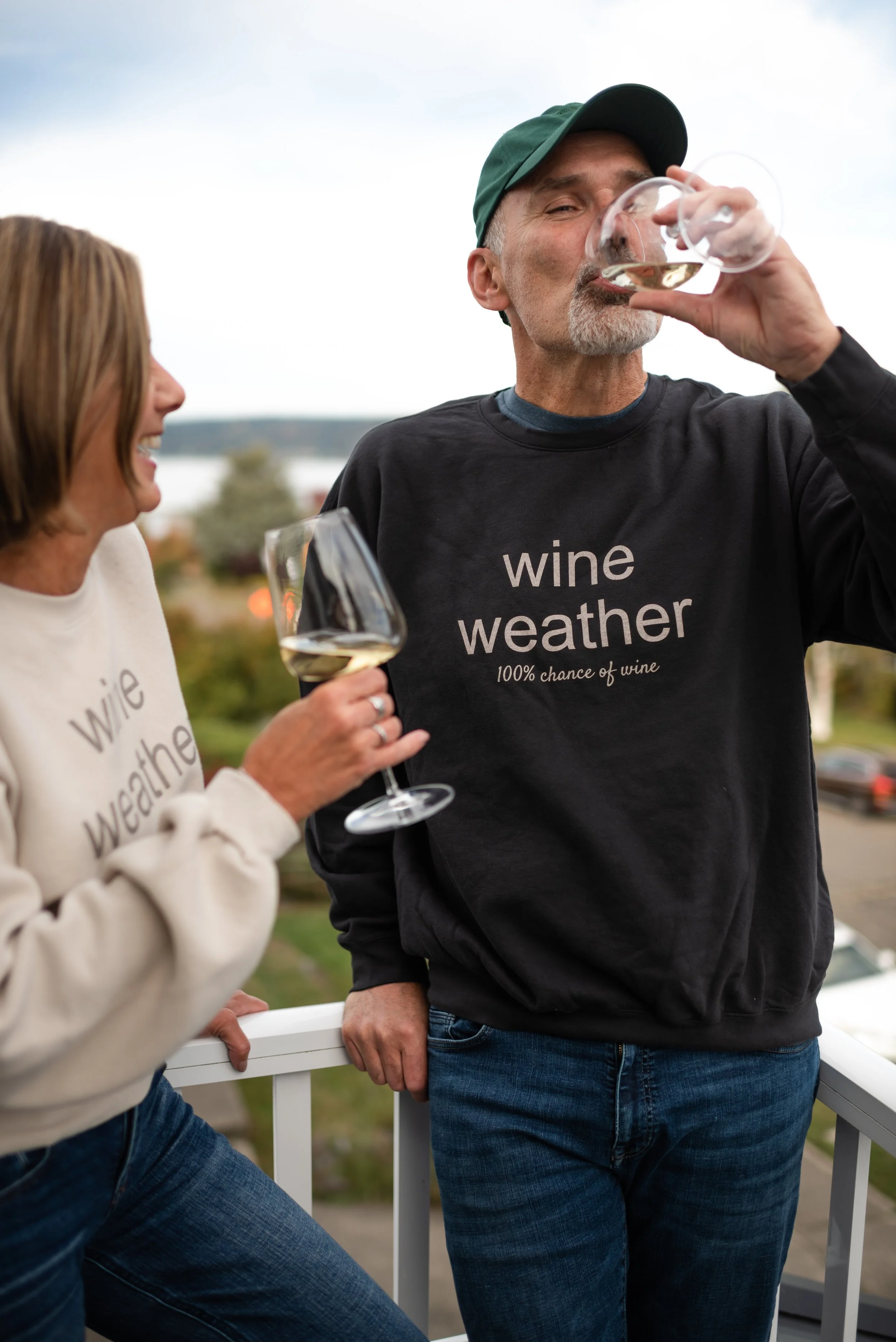 A man and a woman are outdoors on a balcony, holding glasses of white wine and enjoying a drink together. The man is wearing a black sweatshirt with the words "wine weather 100% chance of wine" and the woman is smiling, wearing a cream sweatshirt with the same text. The man is wearing a green cap and is in the process of drinking from his glass, while the woman is looking at him, smiling.