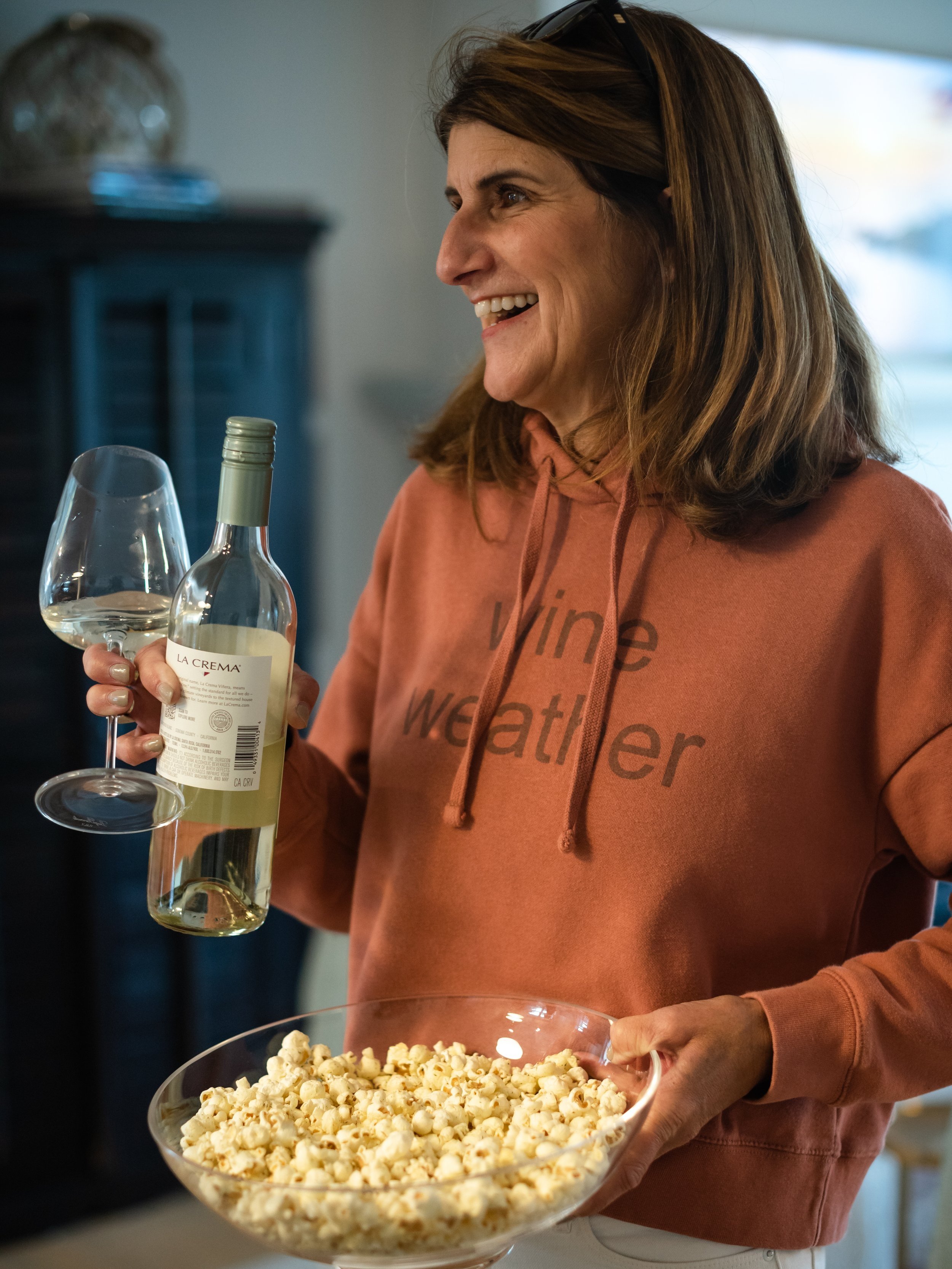 A woman smiling, holding a glass of white wine and a bottle, with a bowl of popcorn on the counter in front of her.