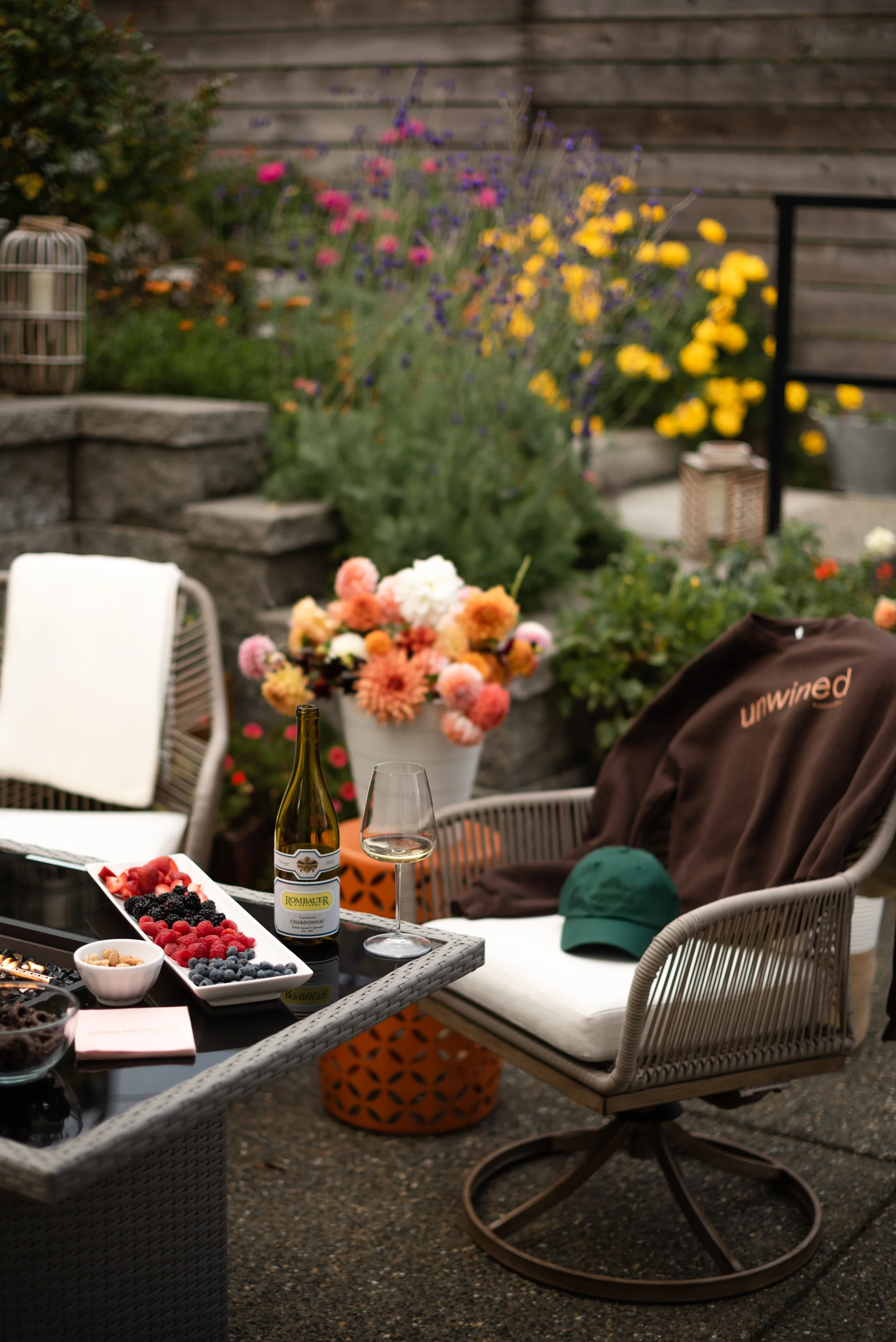 An outdoor patio table with a wine bottle, glass of white wine, and a platter of fresh berries. A cozy wicker chair with a brown blanket and green cap. In the background, colorful flowers and garden plants surround the patio.