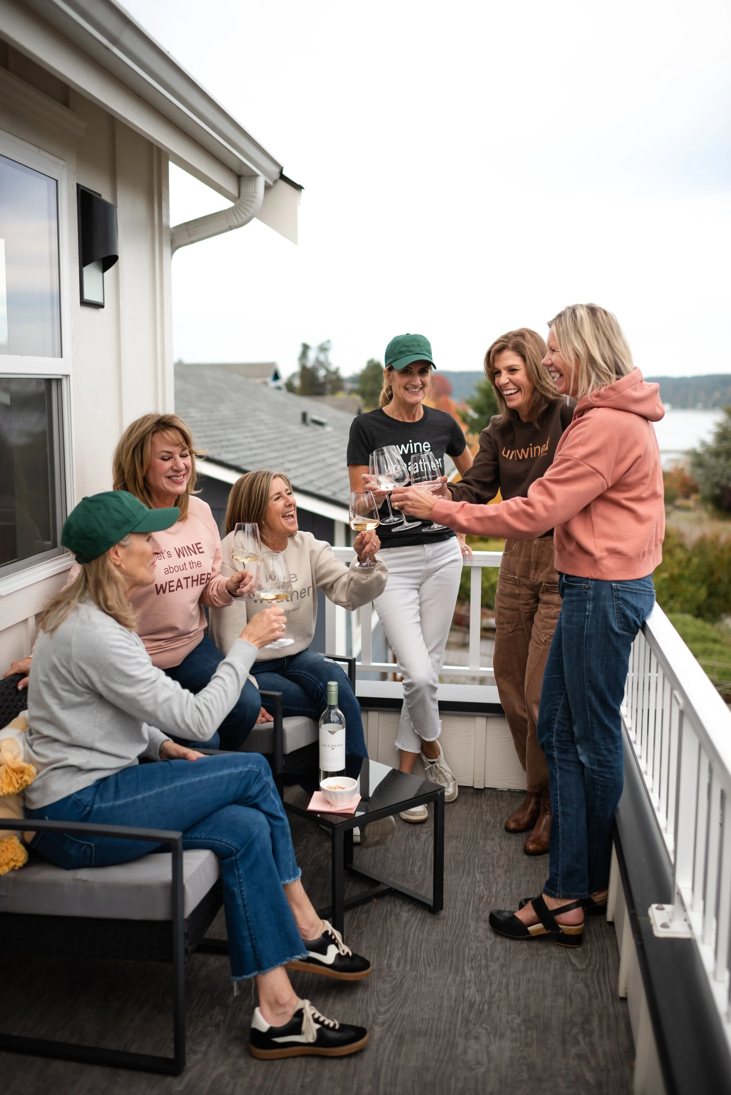 Group of women celebrating on a balcony, holding wine glasses and laughing, with a bottle of wine on the table in front of them.