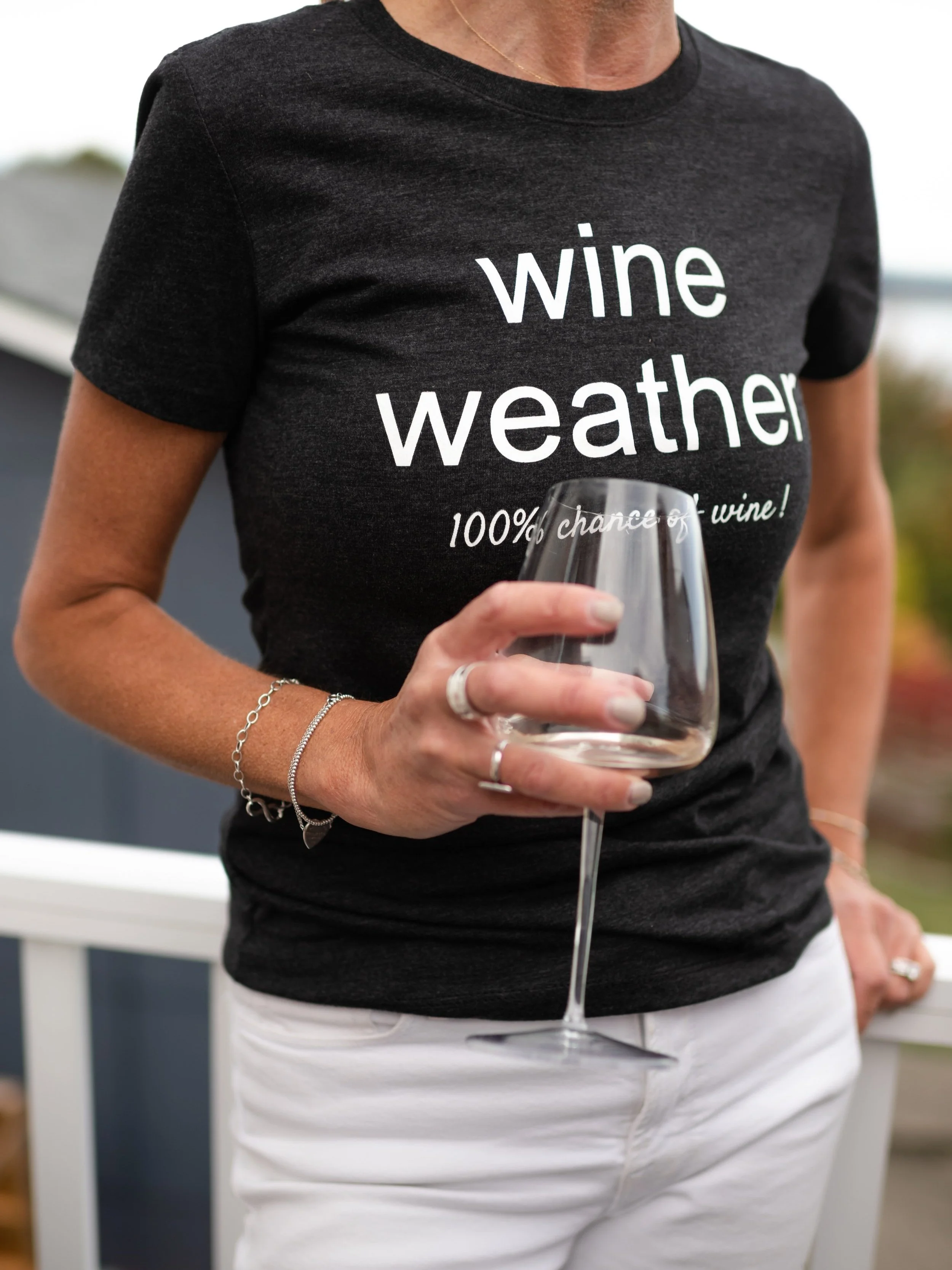 A woman wearing a black T-shirt with white text that says 'wine weather' and '100% chance of wine!' she is holding a wine glass in her right hand, wearing rings and bracelets, standing outdoors with a blurred background.