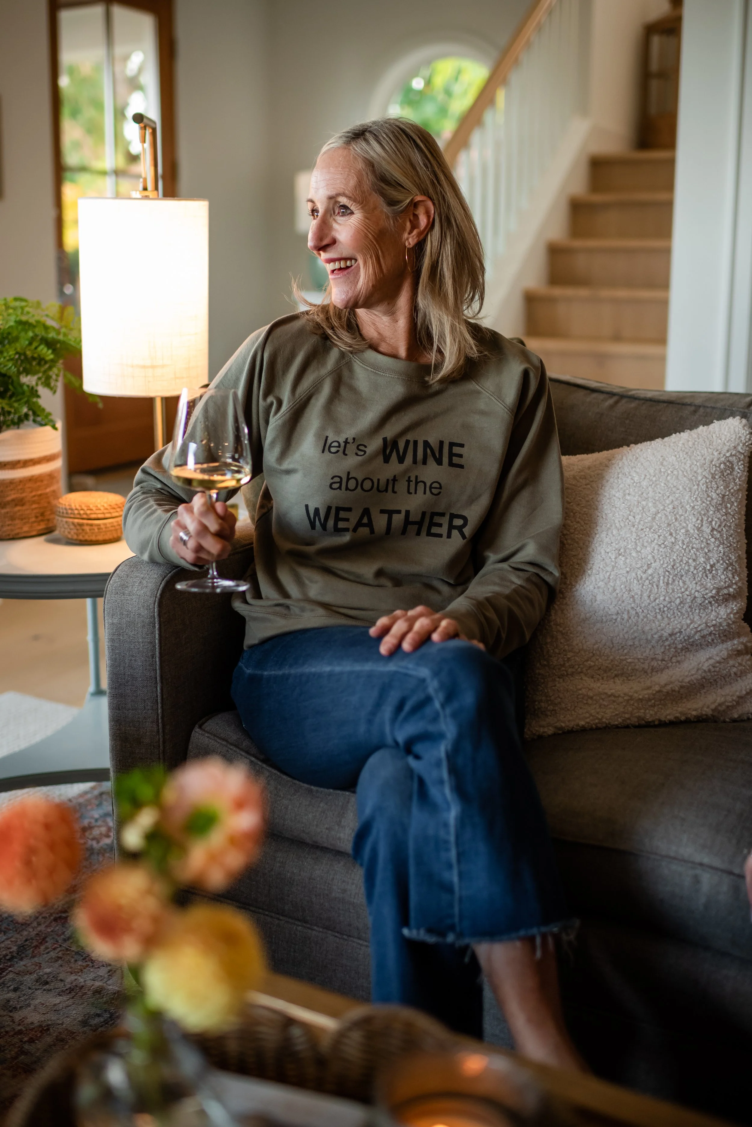 A woman sitting on a gray sofa, smiling, holding a glass of rosé wine, wearing a green sweatshirt reading "let's WINE about the WEATHER."