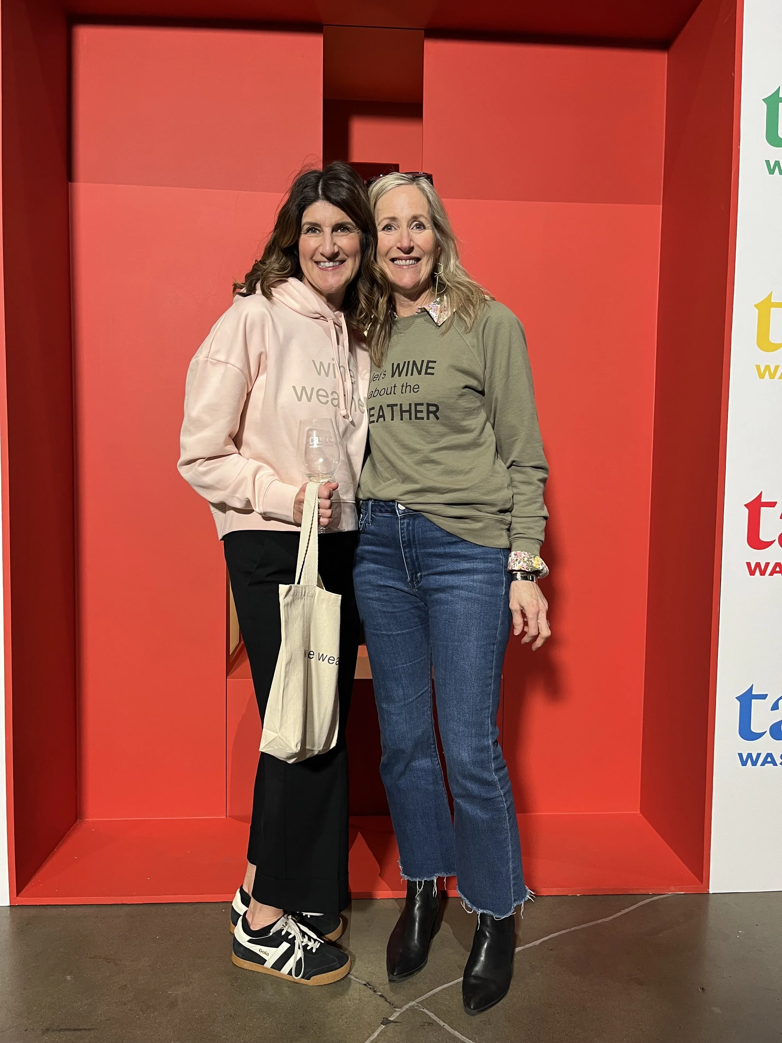 Two women standing close together, smiling at the camera, against a red backdrop. One woman is holding a wine glass and a tote bag. The woman on the left wears a light pink hoodie and black pants, while the woman on the right wears a green sweatshirt and blue jeans.