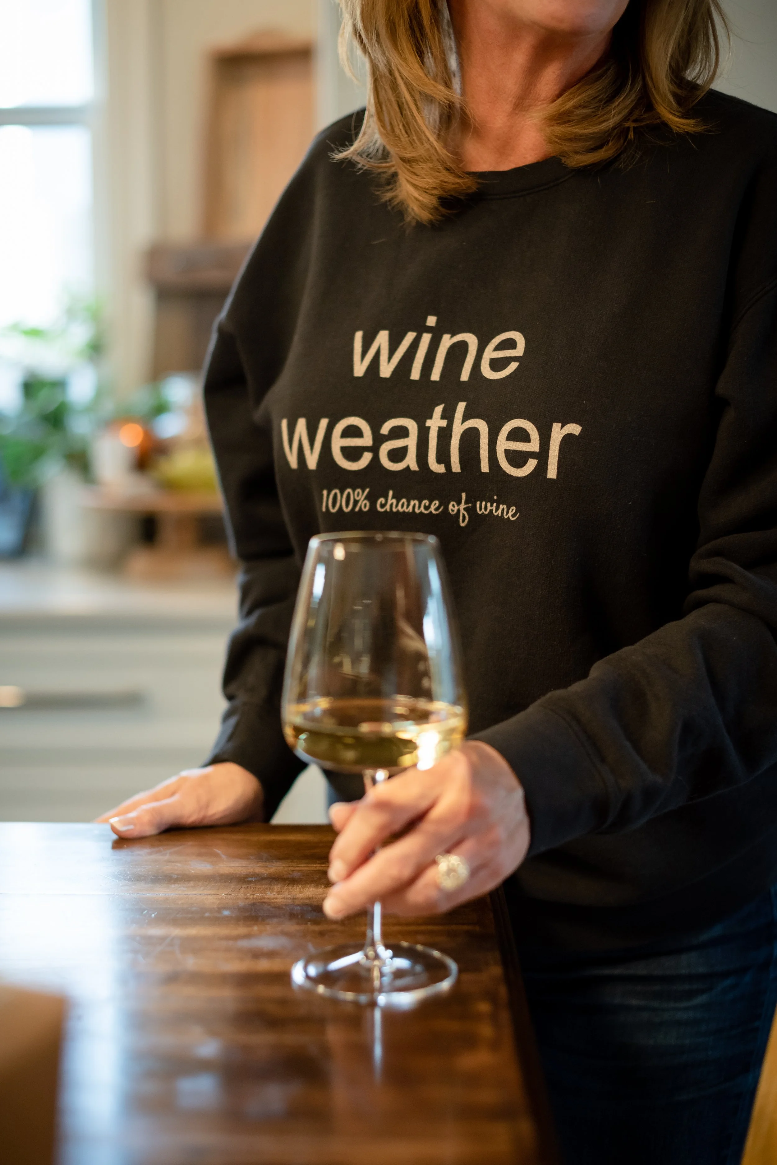 Person wearing a black sweatshirt with 'wine weather 100% chance of wine' written on it, holding a glass of white wine in a kitchen setting.