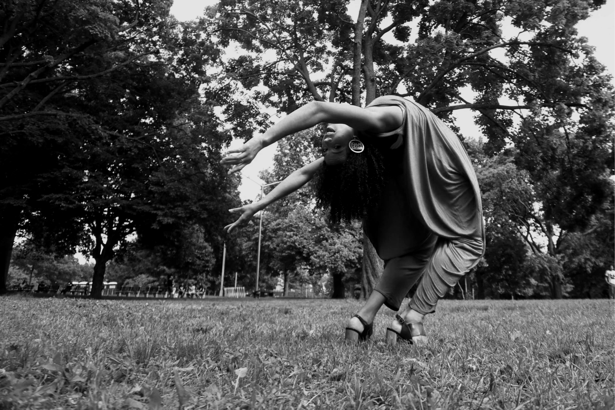 A woman with curly hair poses outdoors on grass, bending backward with arms stretched out, in a Bronx park setting with trees in the background, dressed in a flowing blue jumpsuit and high-heeled shoes.