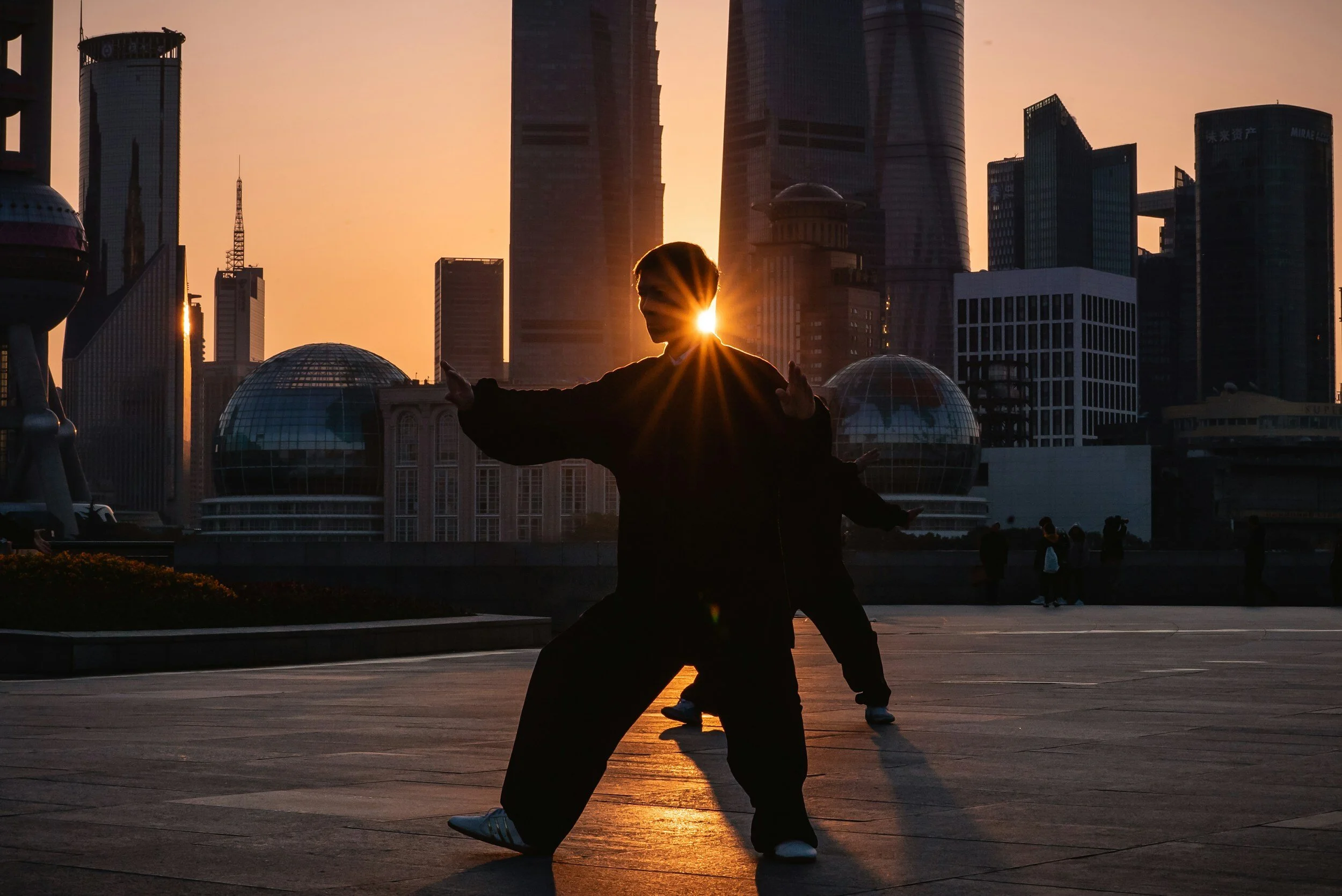 Person practicing martial arts in silhouette at sunrise or sunset in an urban setting with modern skyscrapers and buildings in the background.