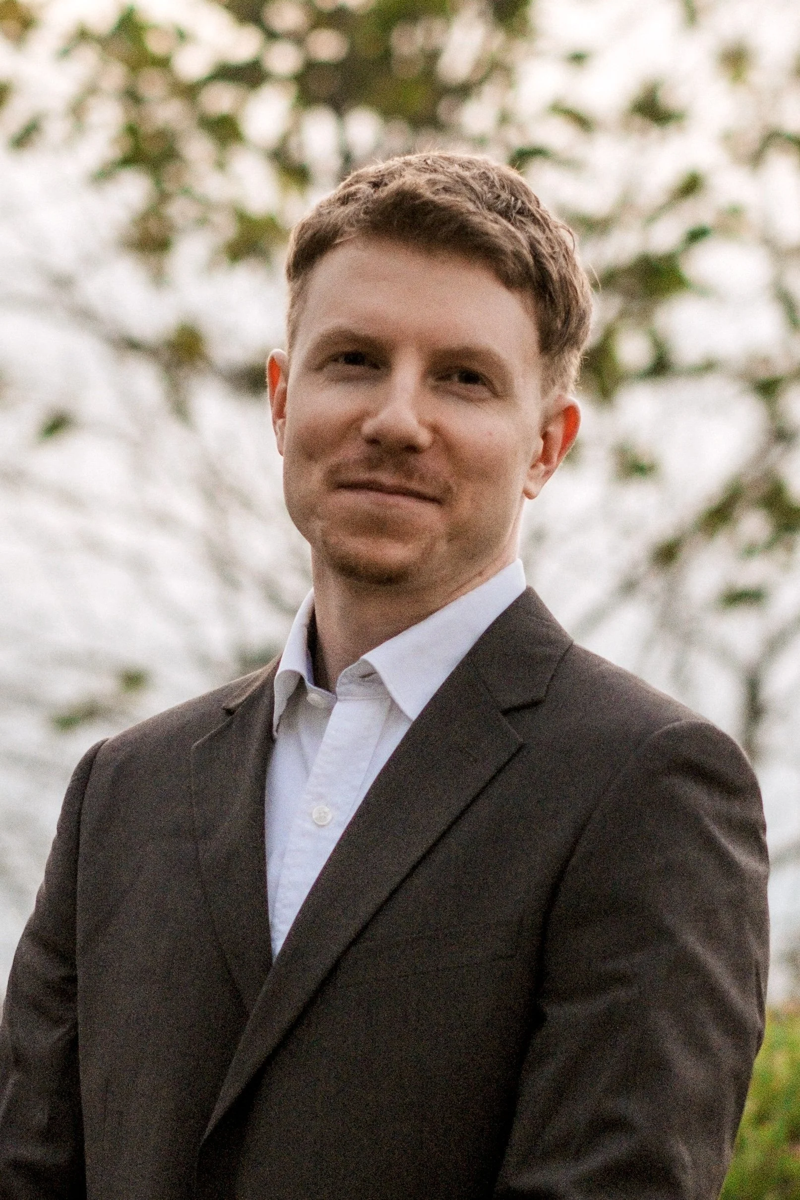 Young man in a suit with white shirt, outdoors, with blurry trees and sky in the background.