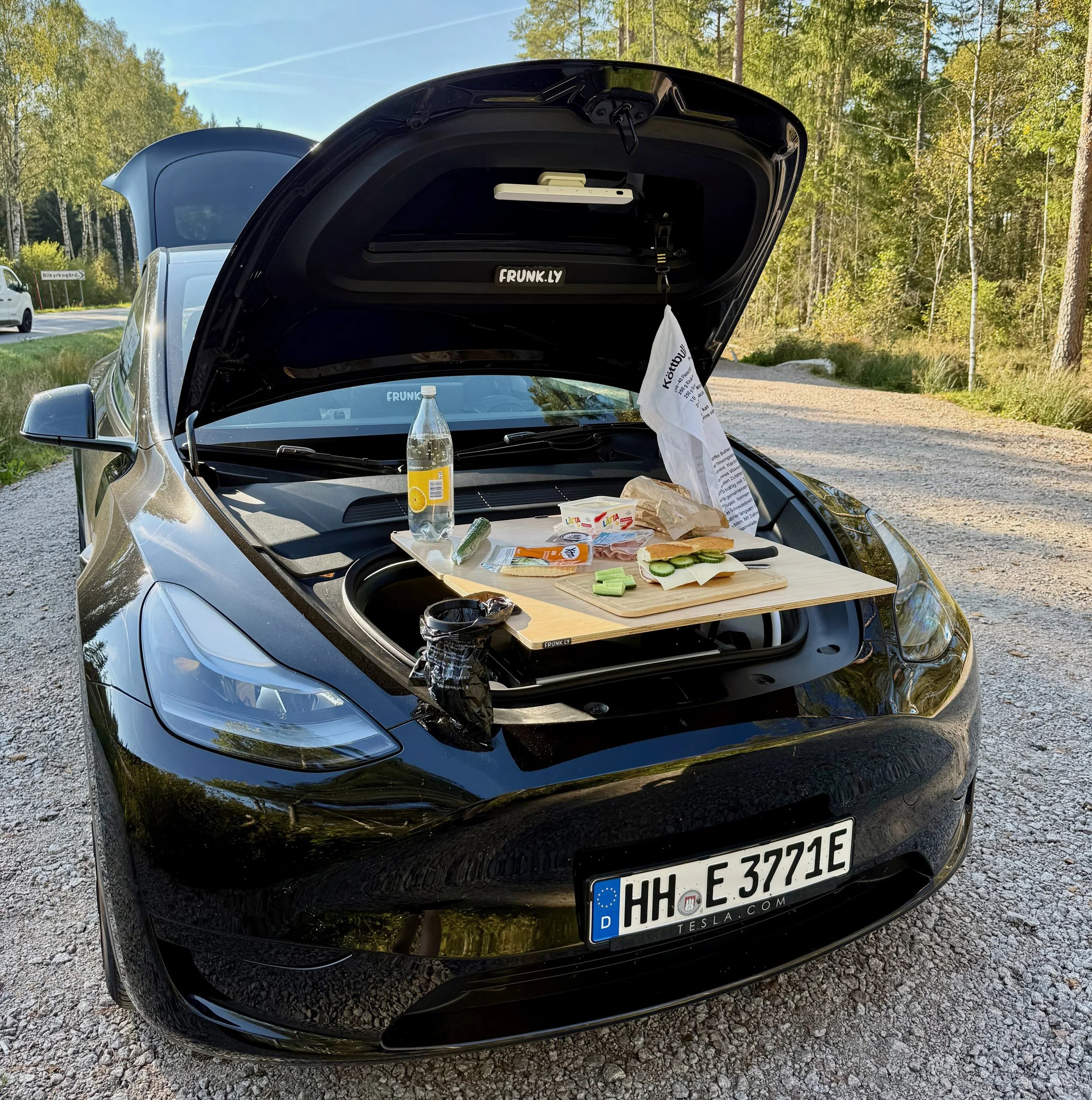 A black Tesla with an open trunk containing a small camping kitchen with food and drinks in a gravel parking lot surrounded by trees.