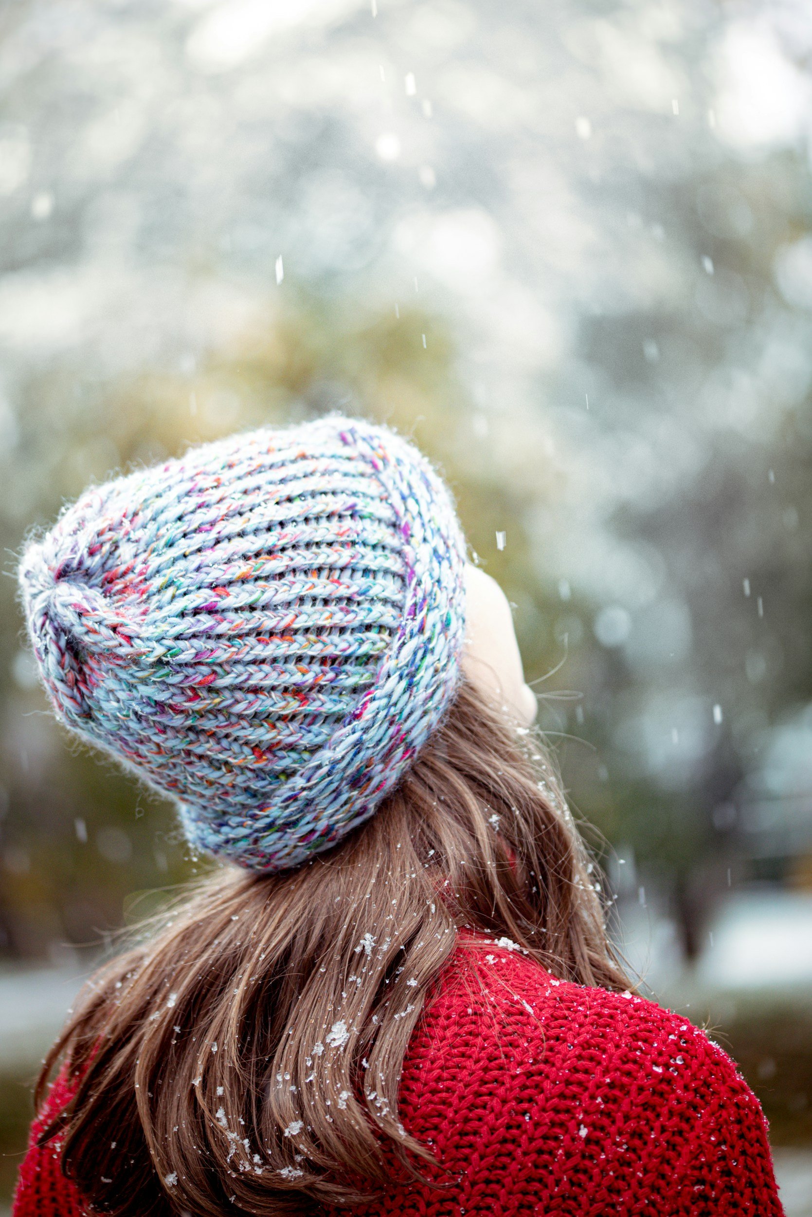 Person wearing a multicolored knit beanie and red sweater, with falling snow and trees in the background.