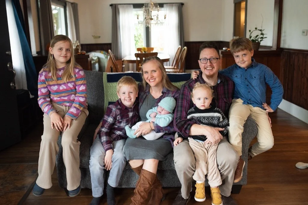 Family of seven sitting on a couch in a living room, smiling, with a dining area in the background.
