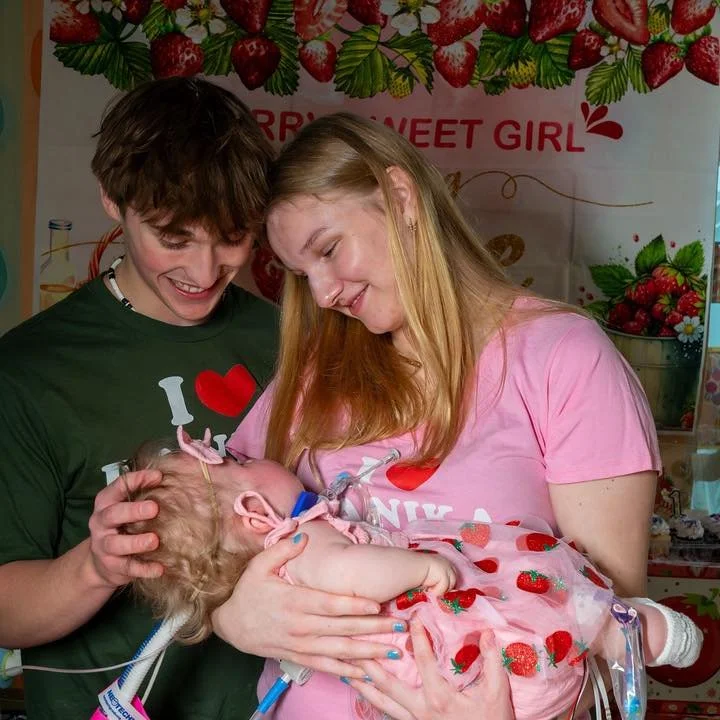 A young couple holding a newborn baby in a hospital setting, all smiling, with a strawberry-themed backdrop.