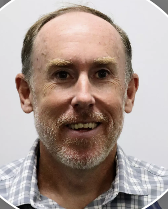 Portrait of a man with light skin, brown hair, wearing a yellow shirt and tie, against a blue background.