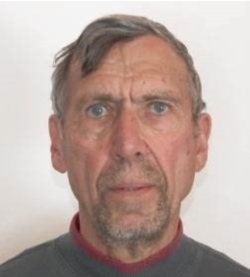 A close-up photo of an elderly man with gray hair, a beard, and missing teeth, wearing a checkered shirt, smiling at the camera.