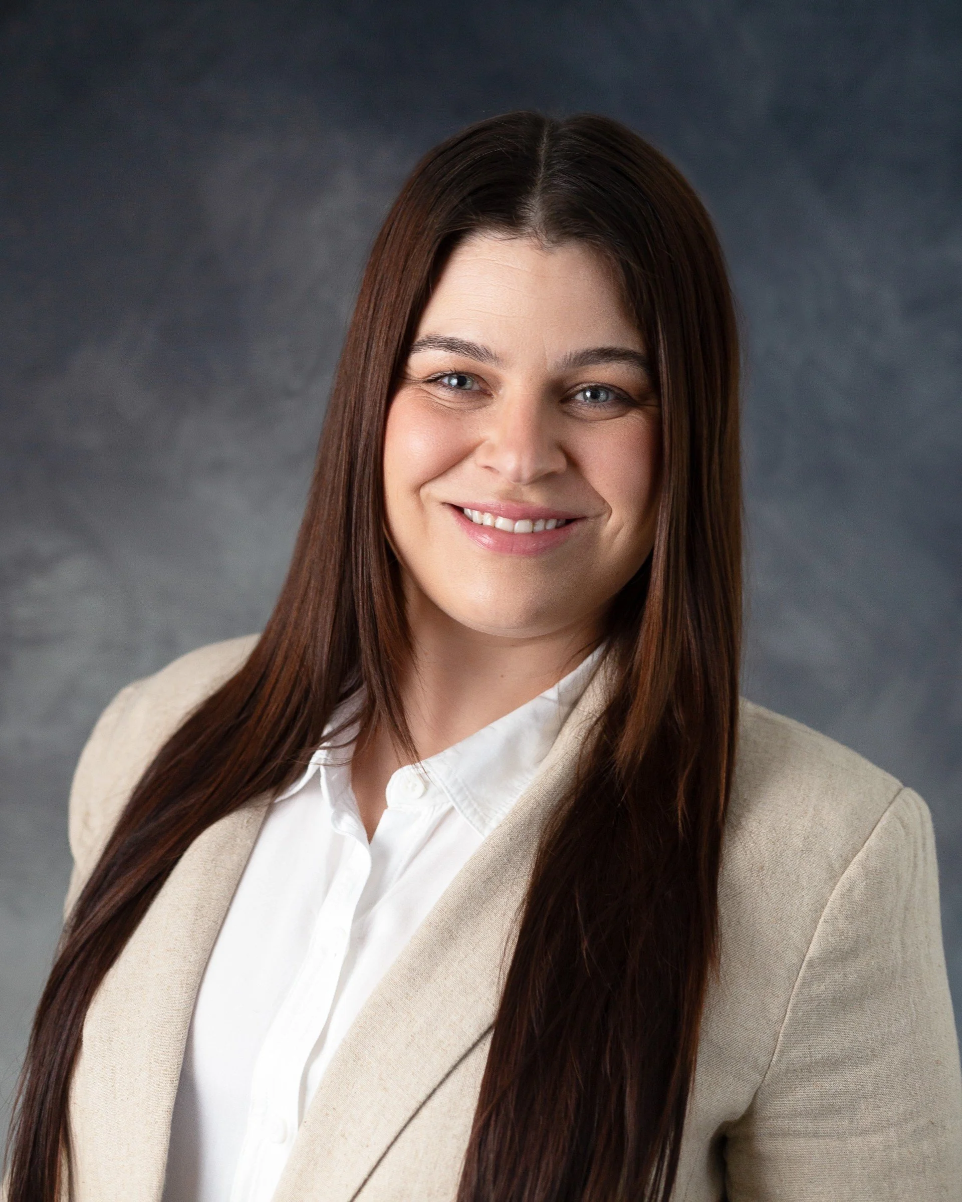 Professional headshot of a woman with long brown hair, wearing a beige blazer over a white blouse, smiling against a dark, textured background.