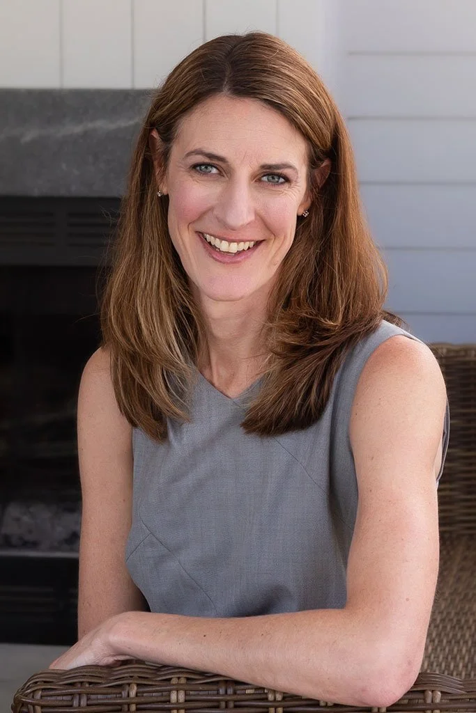 A woman with shoulder-length brown hair and blue eyes, smiling while sitting outdoors in front of a gray wall and a fireplace, wearing a sleeveless gray top.