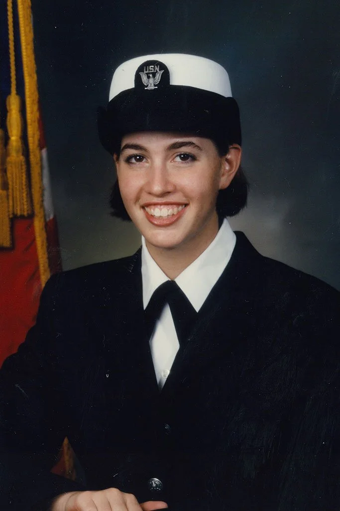 A young woman in U.S. Navy uniform with Rank insignia, wearing a Navy cap with USN emblem, smiling, posed in front of a Navy flag.