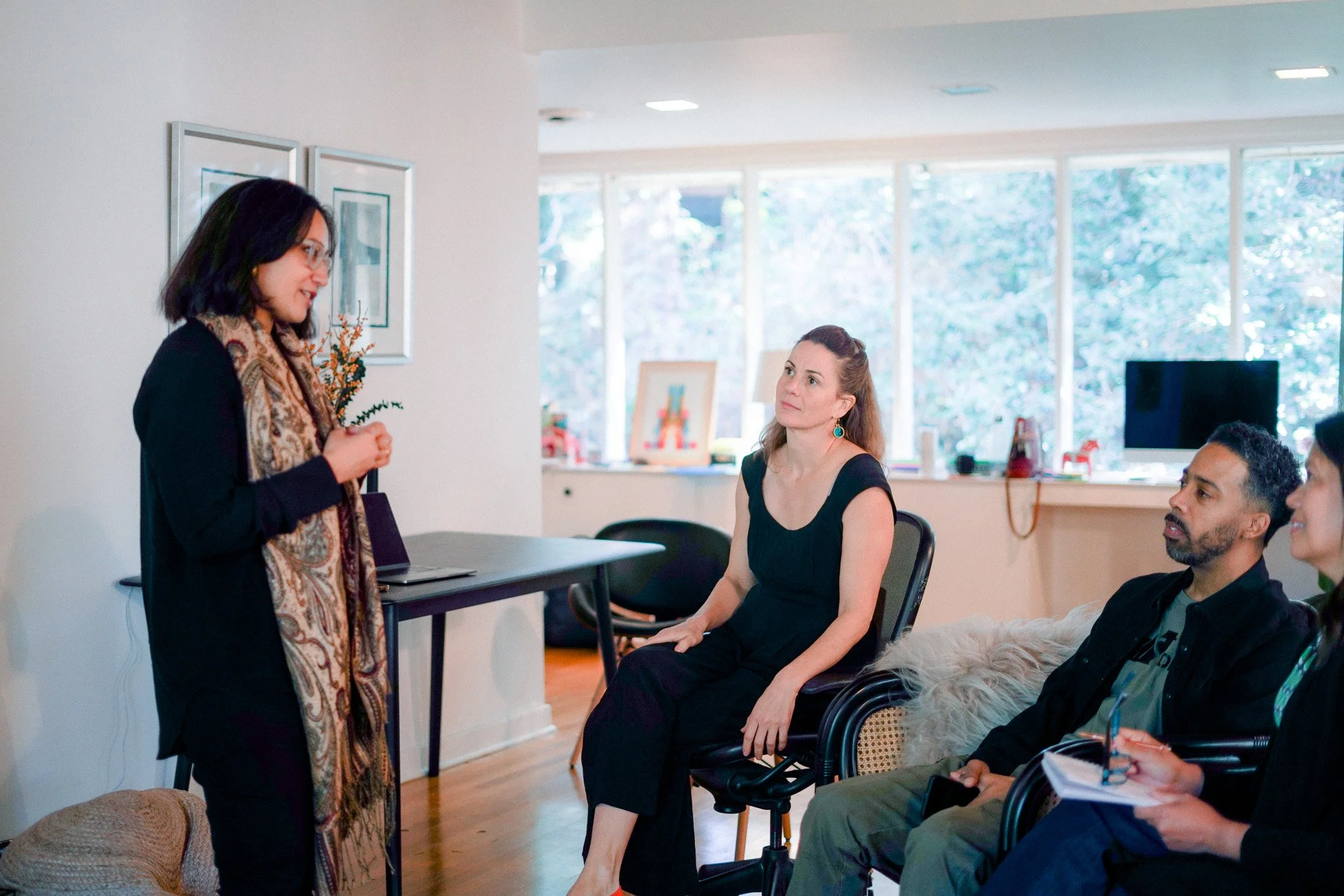 A woman wearing glasses and a patterned scarf is speaking to a group in a modern office setting. Three people are seated, listening attentively. The room is bright with large windows and a desk in the background.