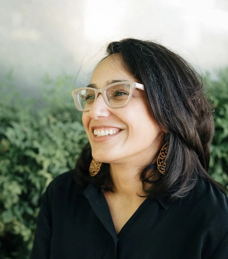 Smiling person with glasses and long hair wearing a black shirt, standing outdoors with greenery in the background.