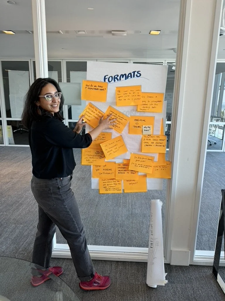 Person standing next to a board with "FORMATS" written at the top, covered with orange sticky notes containing handwritten text.
