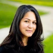 Portrait of a woman with long dark hair and a neutral expression, standing outdoors with a blurred grassy background.