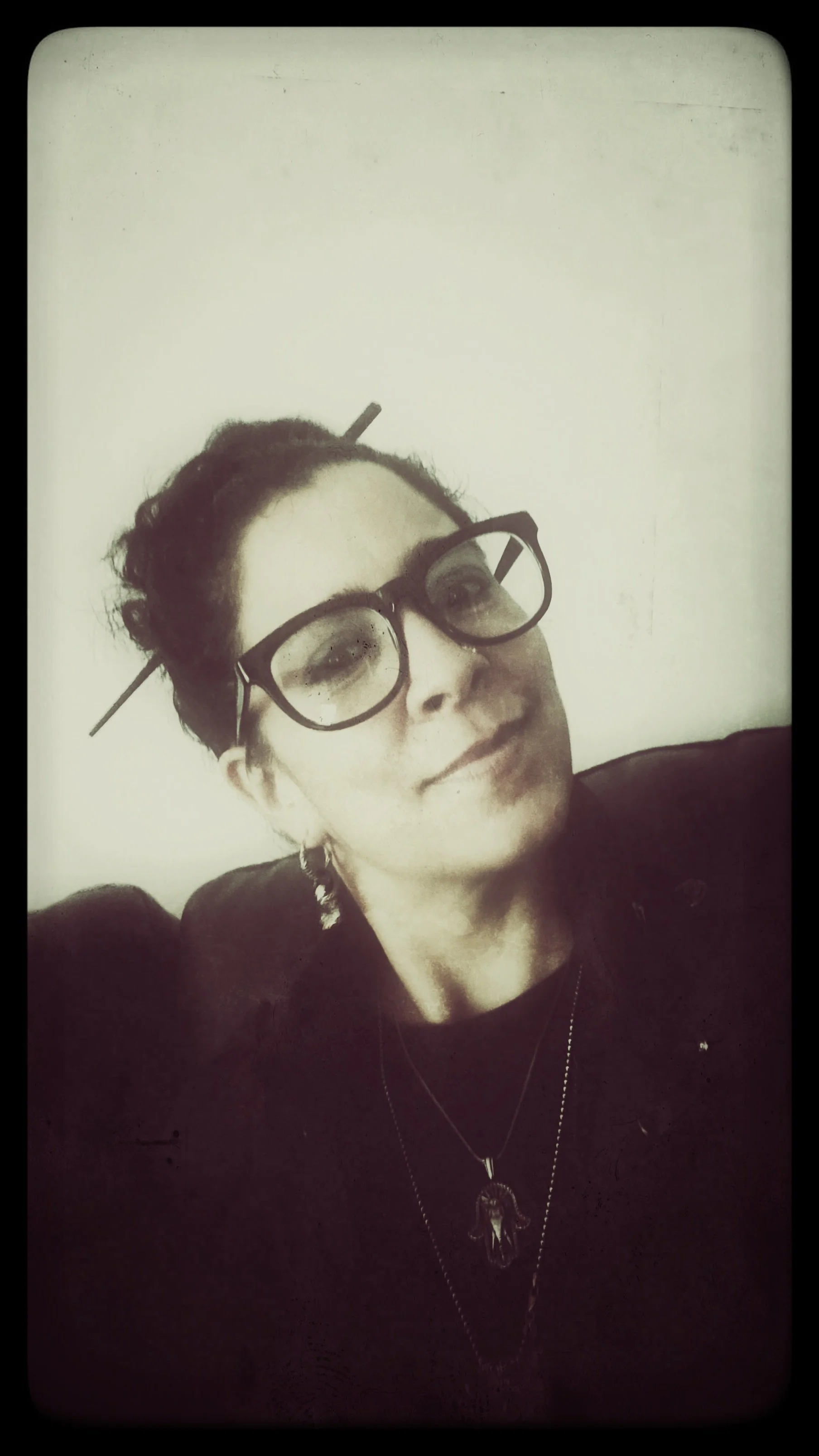 Person wearing glasses with hair tied up, holding a stick in hair, necklace, and earrings. Sepia tone photograph.