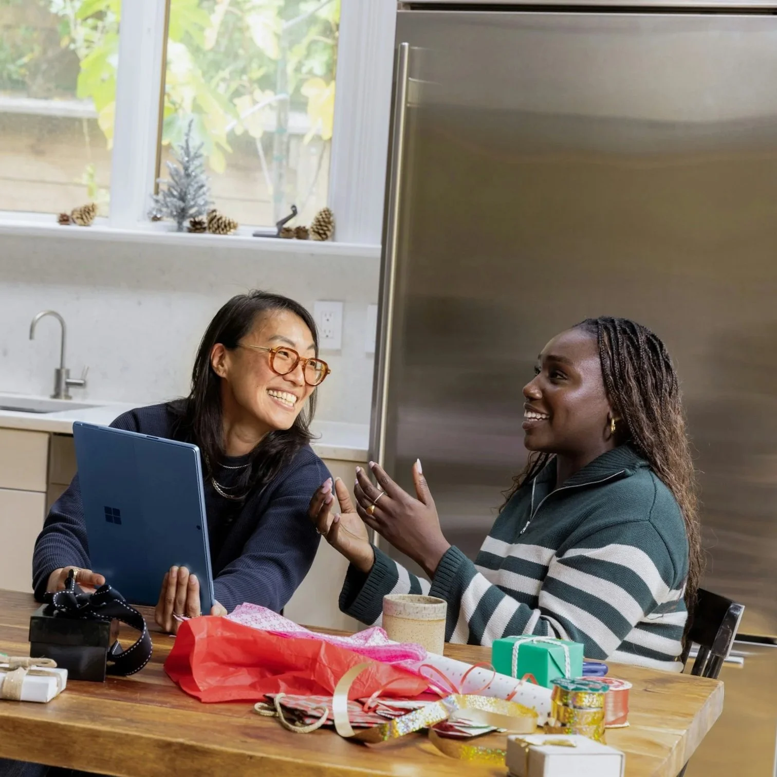 Two women are sitting at a table in a kitchen, smiling and engaging in conversation. The table has wrapping paper, ribbons, and small gift boxes, suggesting a gift-giving occasion.