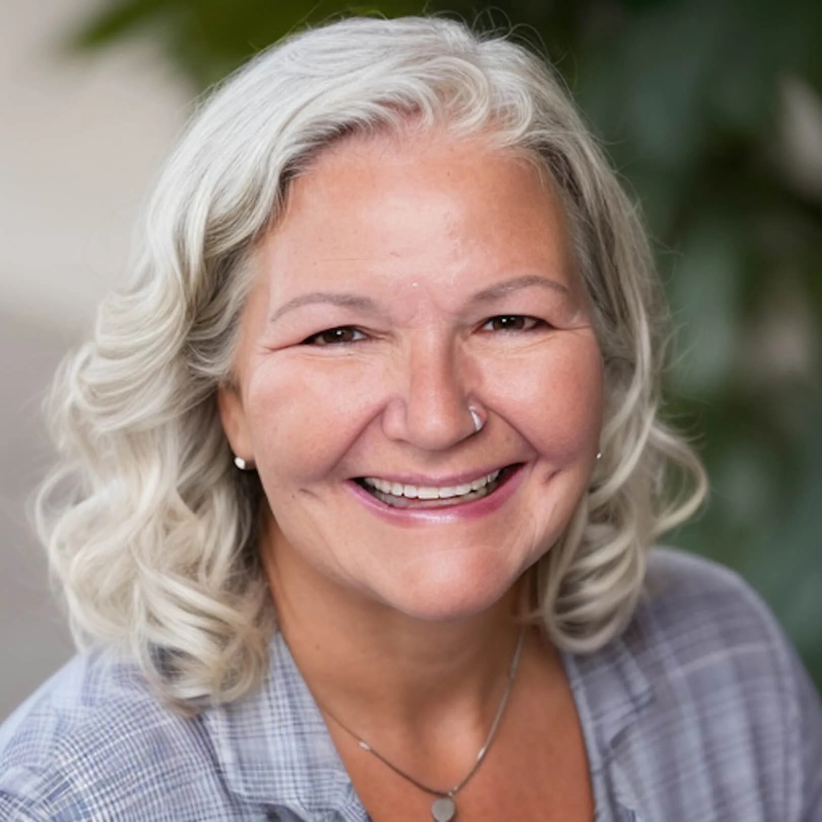 Close-up of a smiling woman with shoulder-length wavy gray hair, wearing a checkered shirt, small earrings, and a necklace with a pendant.