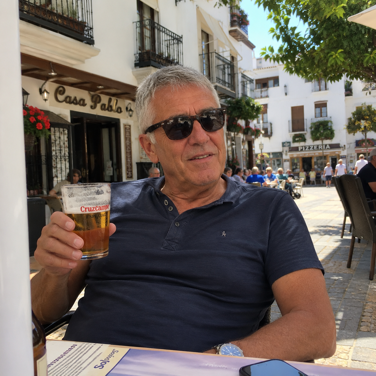 A man wearing sunglasses and a navy polo shirt, sitting at an outdoor café, holding a glass of beer with the label 'Cruzcampo,' smiling, with a bustling square, pizzeria, and white buildings with balconies in the background under a sunny sky.