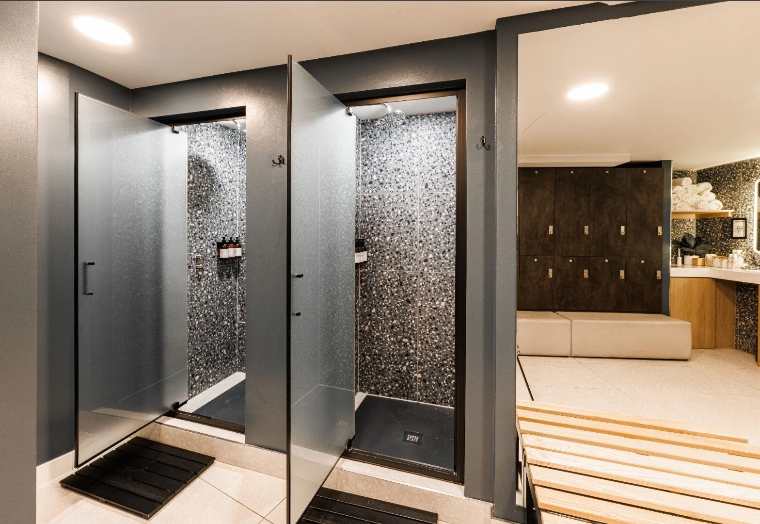 Two shower stalls with frosted glass doors and black speckled tile walls, one with an overhead shower. To the right, a wooden cabinet with lockers and a seating area, with a vanity and towels in the background.