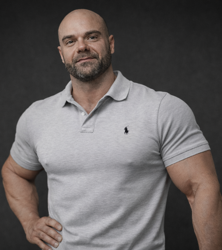 A bald man with a beard, wearing a light gray polo shirt with a small black logo, standing confidently with one hand on his hip against a dark background.