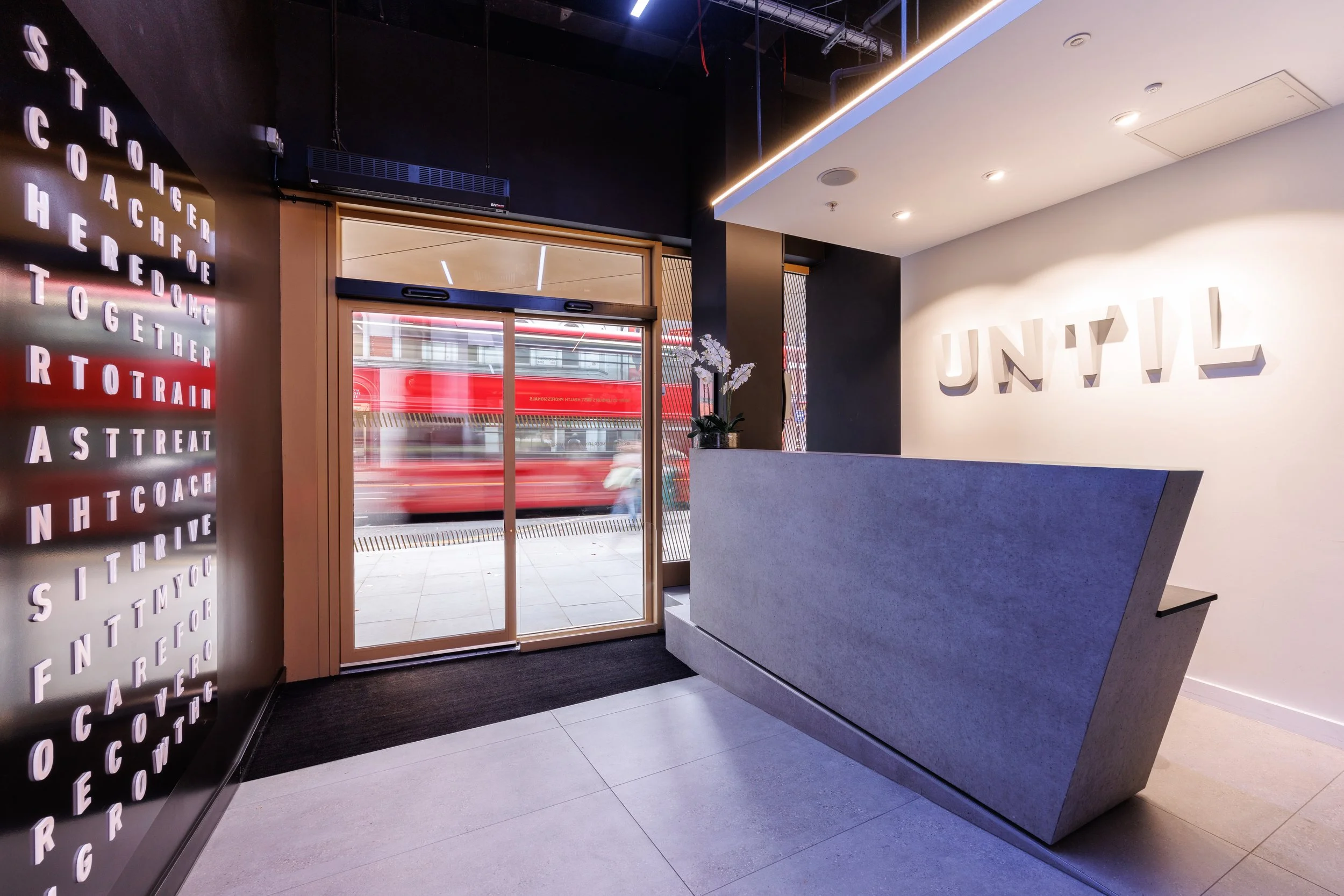 Modern interior of a building lobby with a sleek reception desk, large wall sign with the word 'UNLY', an eye-catching letterboard with words like 'STROIGE', 'COACH', and 'THRIVE', large glass sliding doors leading outside, and a view of a red double