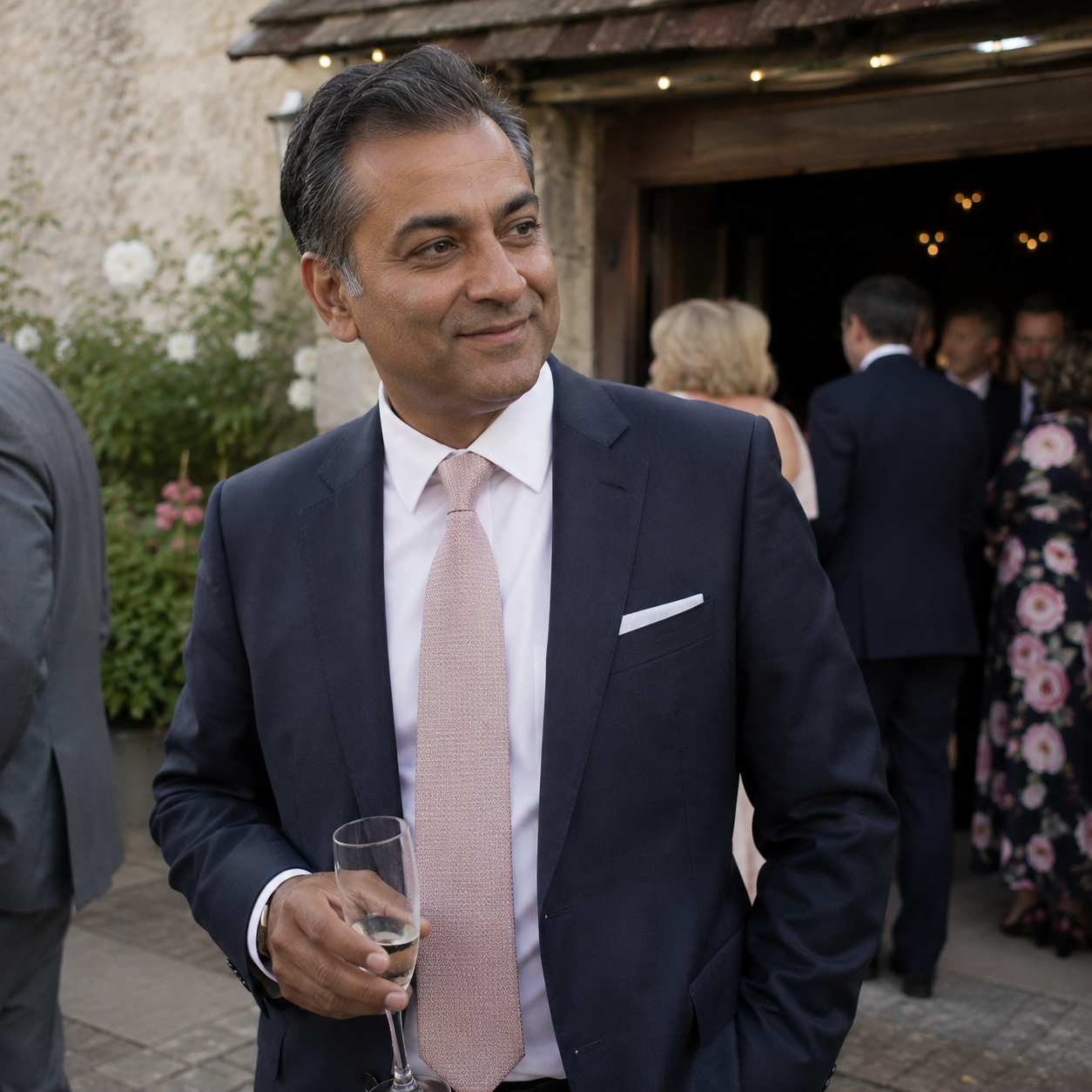 A man in a dark suit and pink tie holding a champagne flute at a social gathering outside, with other guests in formal attire in the background.