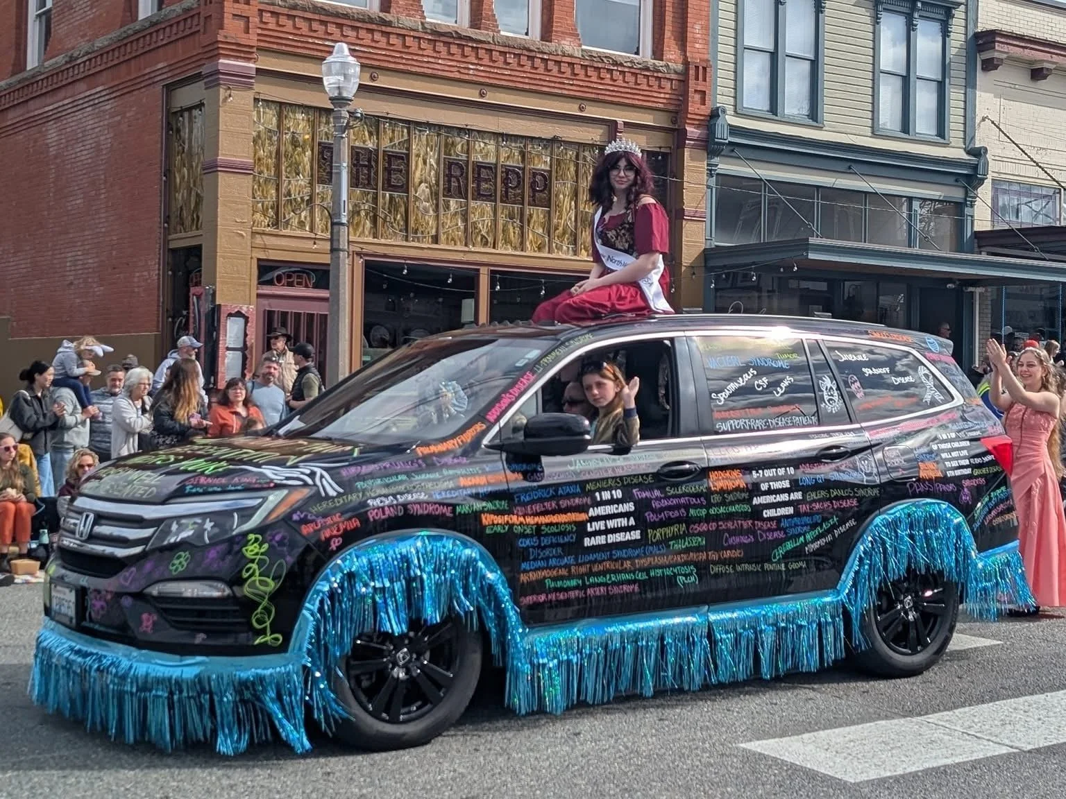 Giusiana Prosser Rides in the Kla Ha Ya Days Easter Parade on top of a black SUV covered in colorful rare disease names