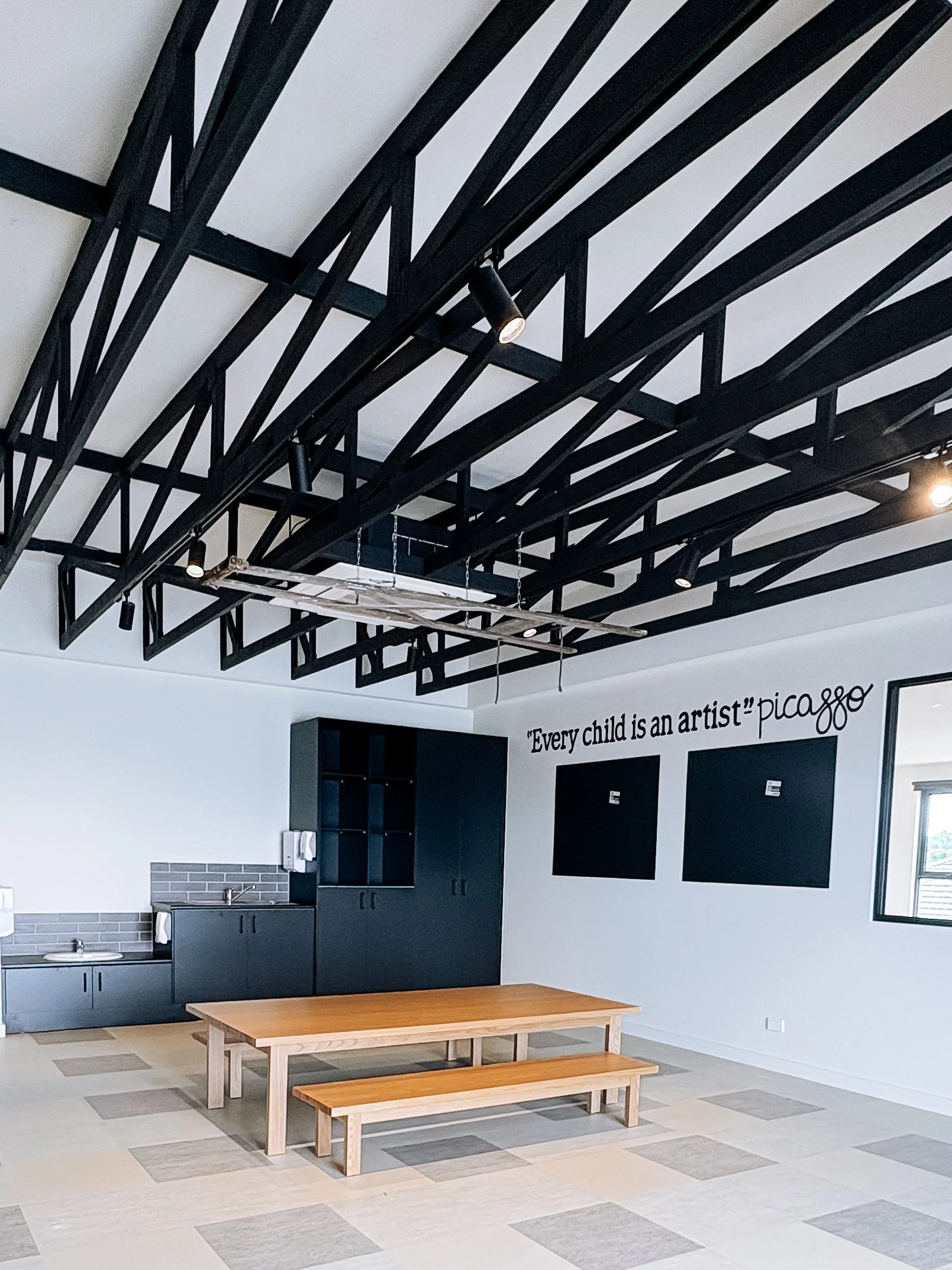 Empty classroom or activity room with a wooden table and bench, black cabinets and wall chalkboards, quote on wall saying "Every child is an artist" Picasso, and a window, with exposed black ceiling beams and cloudy sky outside.