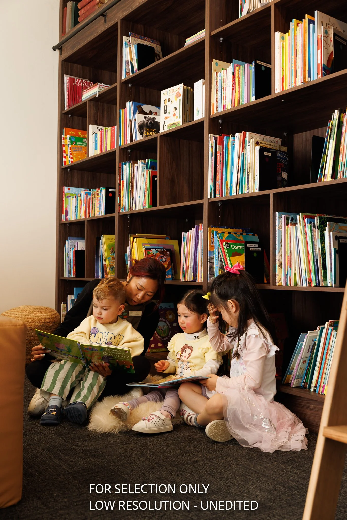 A woman reading a book with three young children in a cozy library corner, sitting on a plush rug in front of a large bookshelf filled with colorful children's books.