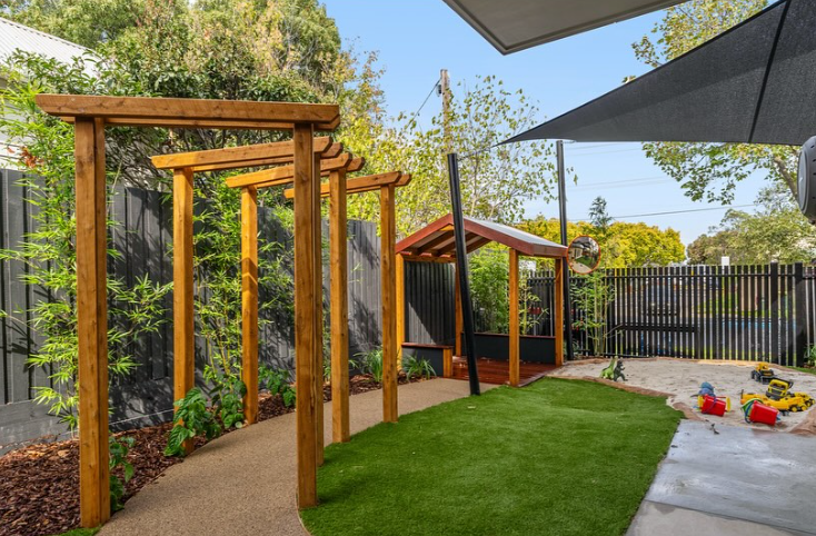 Backyard with a wooden swing set frame, small playhouse, artificial grass, sand area with toys, shaded by an umbrella, enclosed with a black fence, and surrounded by trees.