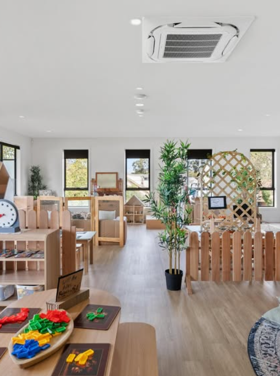 A brightly lit classroom with wooden furniture, green plants, and windows with black frames, creating a welcoming learning environment.