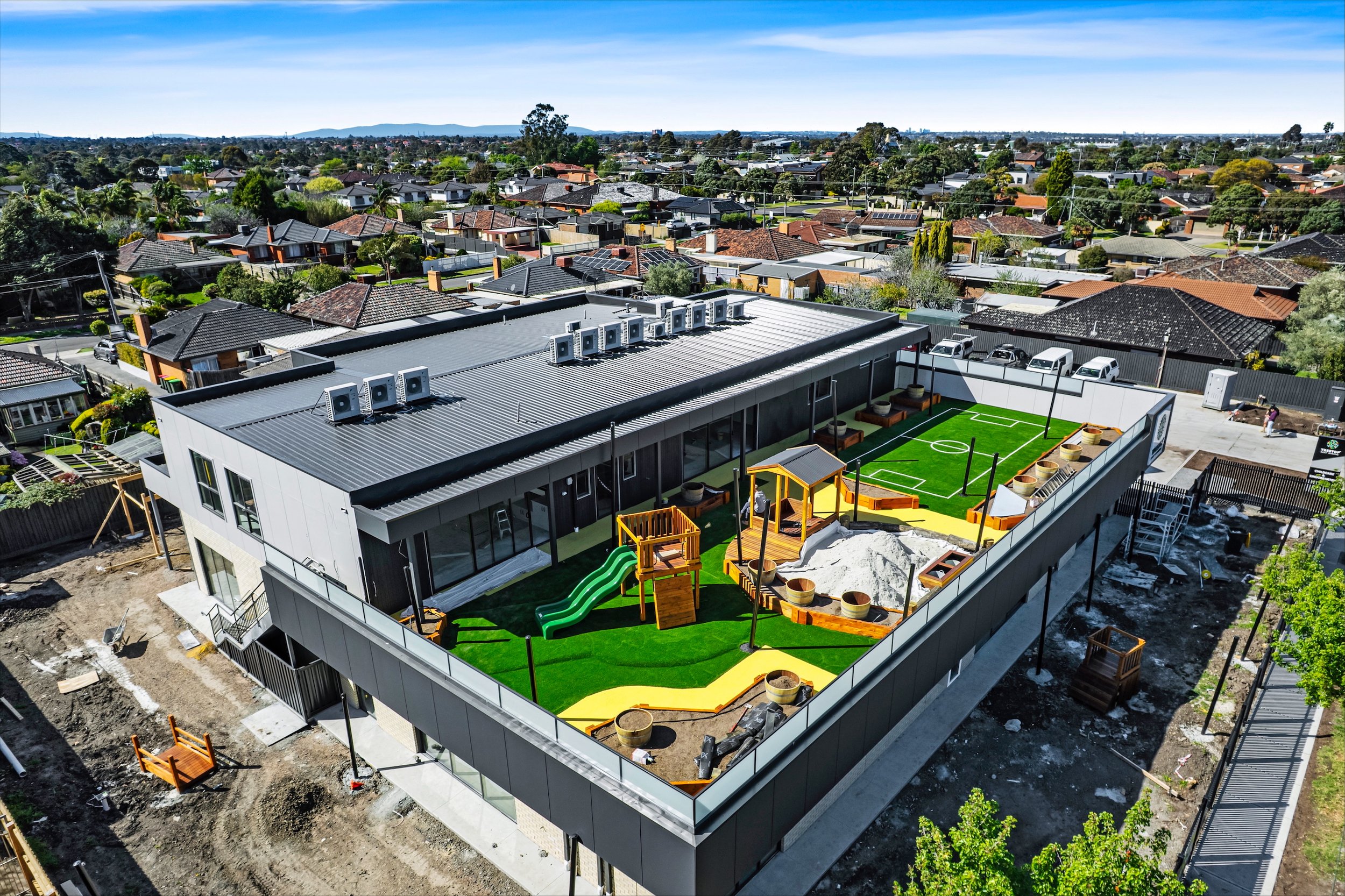 A modern building with a rooftop playground and sports area, including a small soccer field, a slide, sandbox, and seating areas, located in a residential neighborhood.