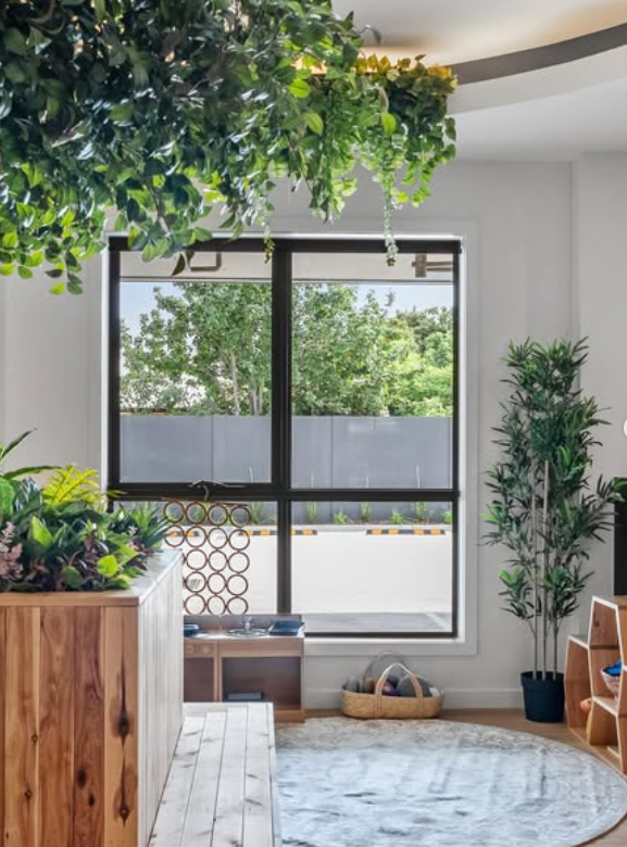 Interior room with large window, green plants, wooden furniture, and a round rug, with trees visible outside.