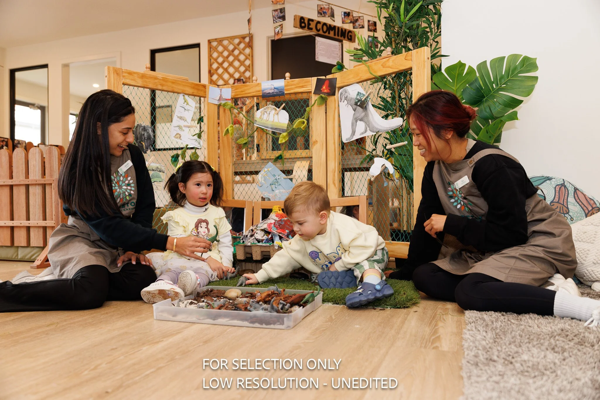 Two teachers and two young children sitting on the floor in a classroom, engaging with toy dinosaurs in a tray, with educational dinosaur pictures hanging on a wooden and wire divider behind them.