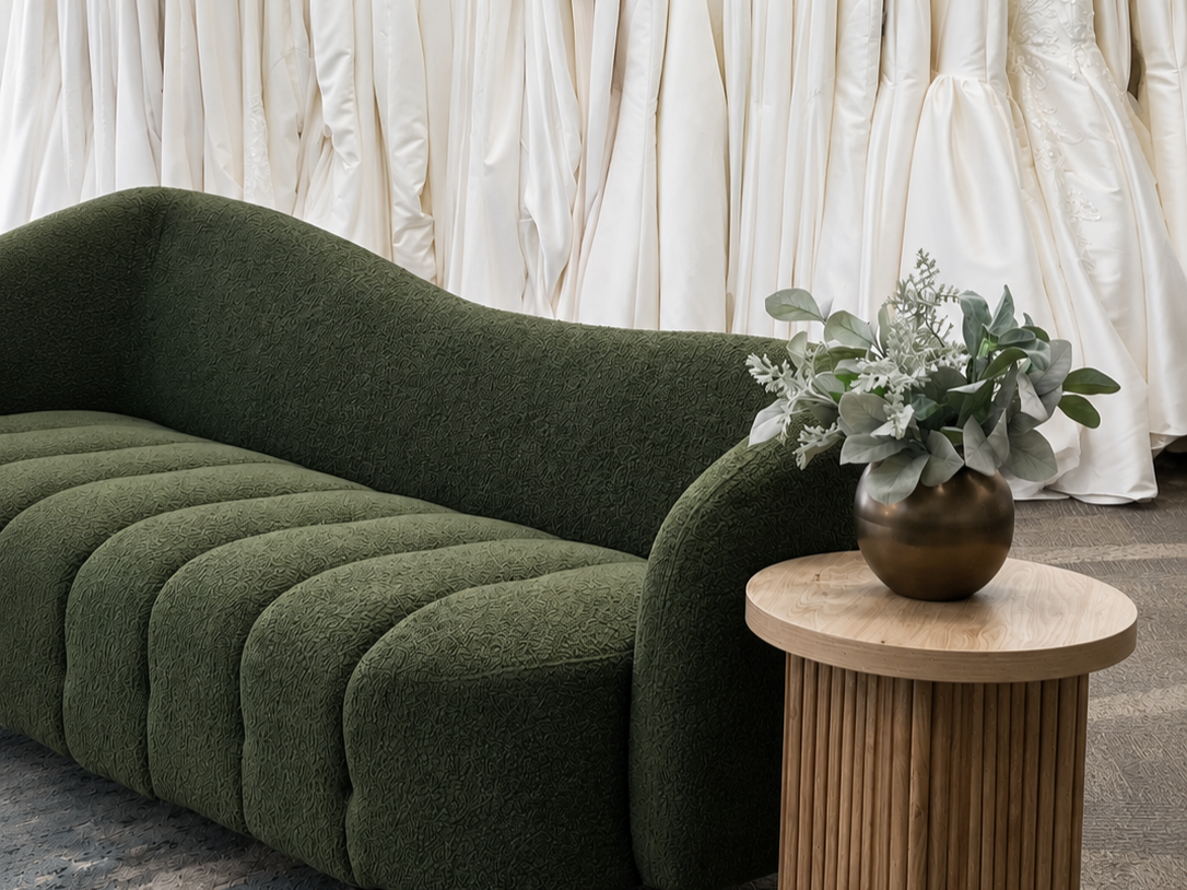 A green upholstered sofa with a curved backrest and textured fabric, next to a round wooden side table with a dark vase holding white and gray artificial flowers, in front of a backdrop of white draped fabric.