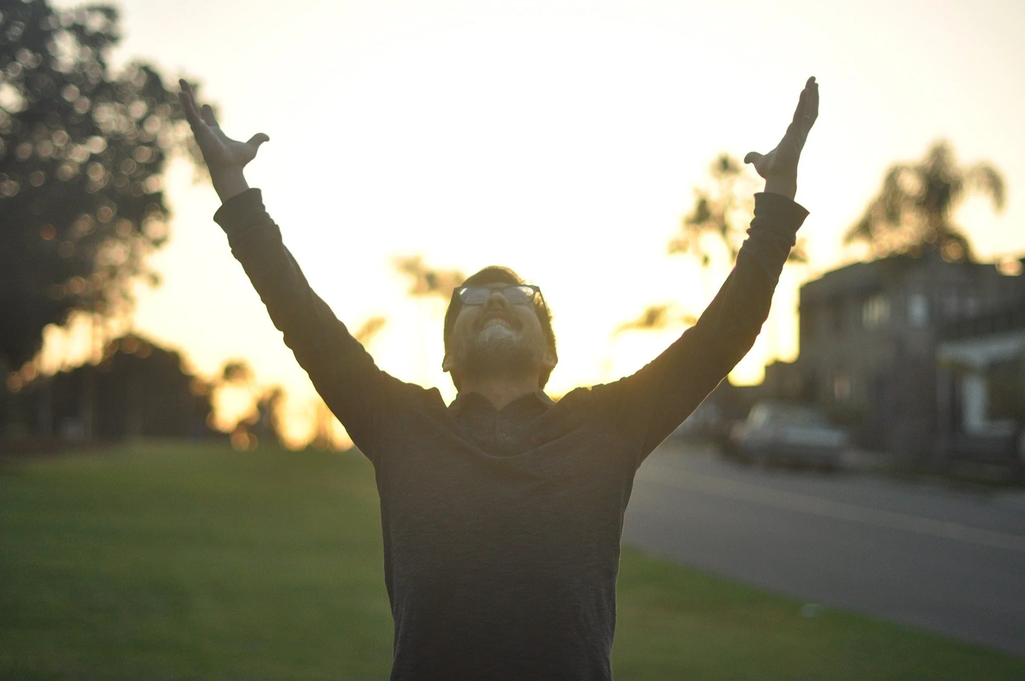 Chiropractor Koichi Naruishi stands facing the camera with his arms stretch upwards with the sun behind him in Encinitas, California