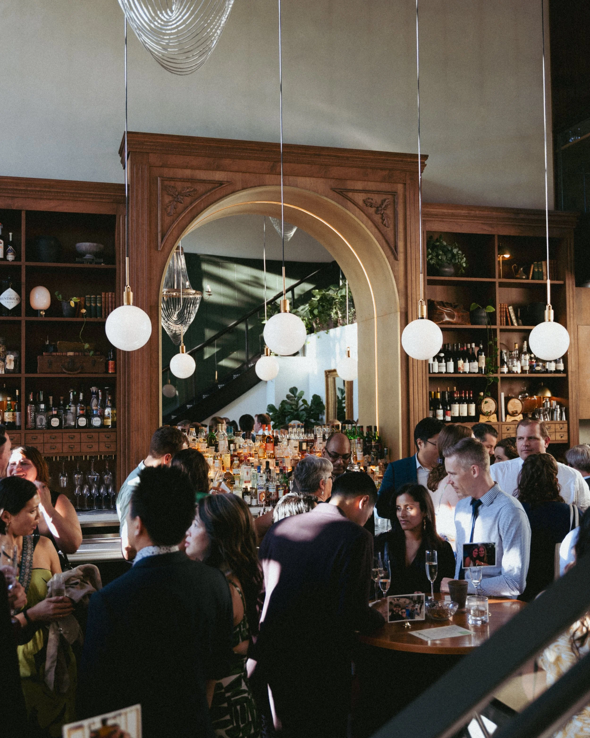 People socializing inside a bar or restaurant with wood shelving, bottles, and a large mirror.