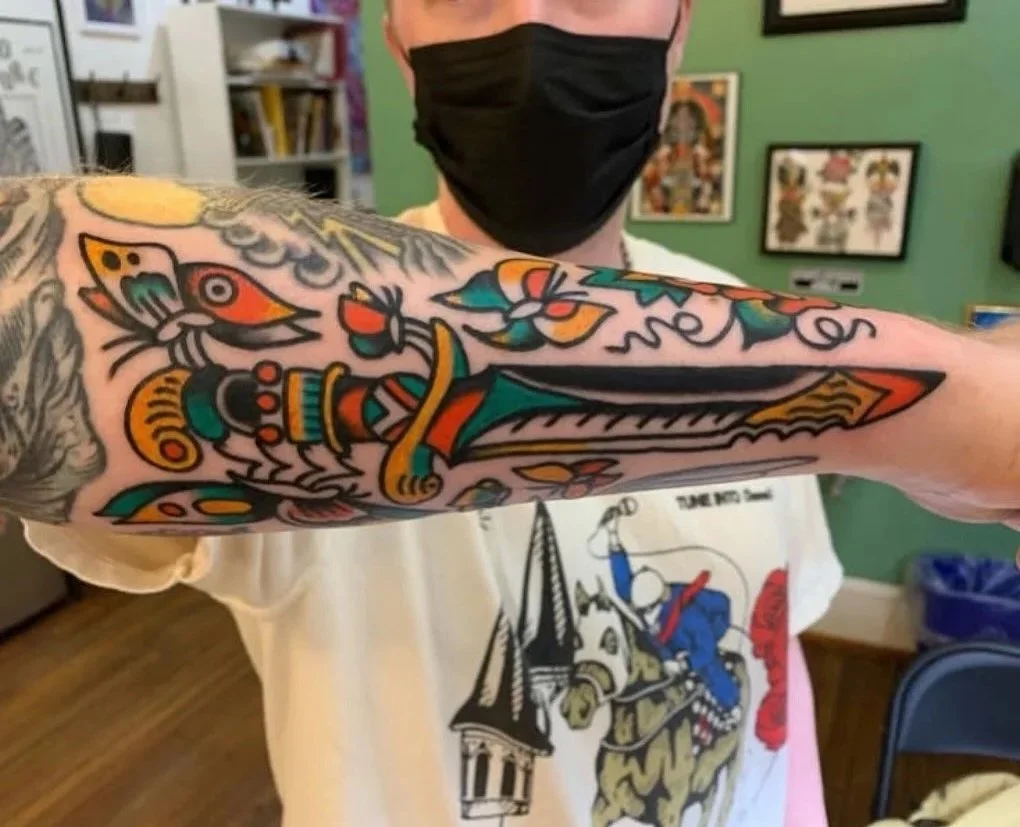 Colorful traditional tattoo featuring a dagger, butterflies, and floral designs on a person's forearm.