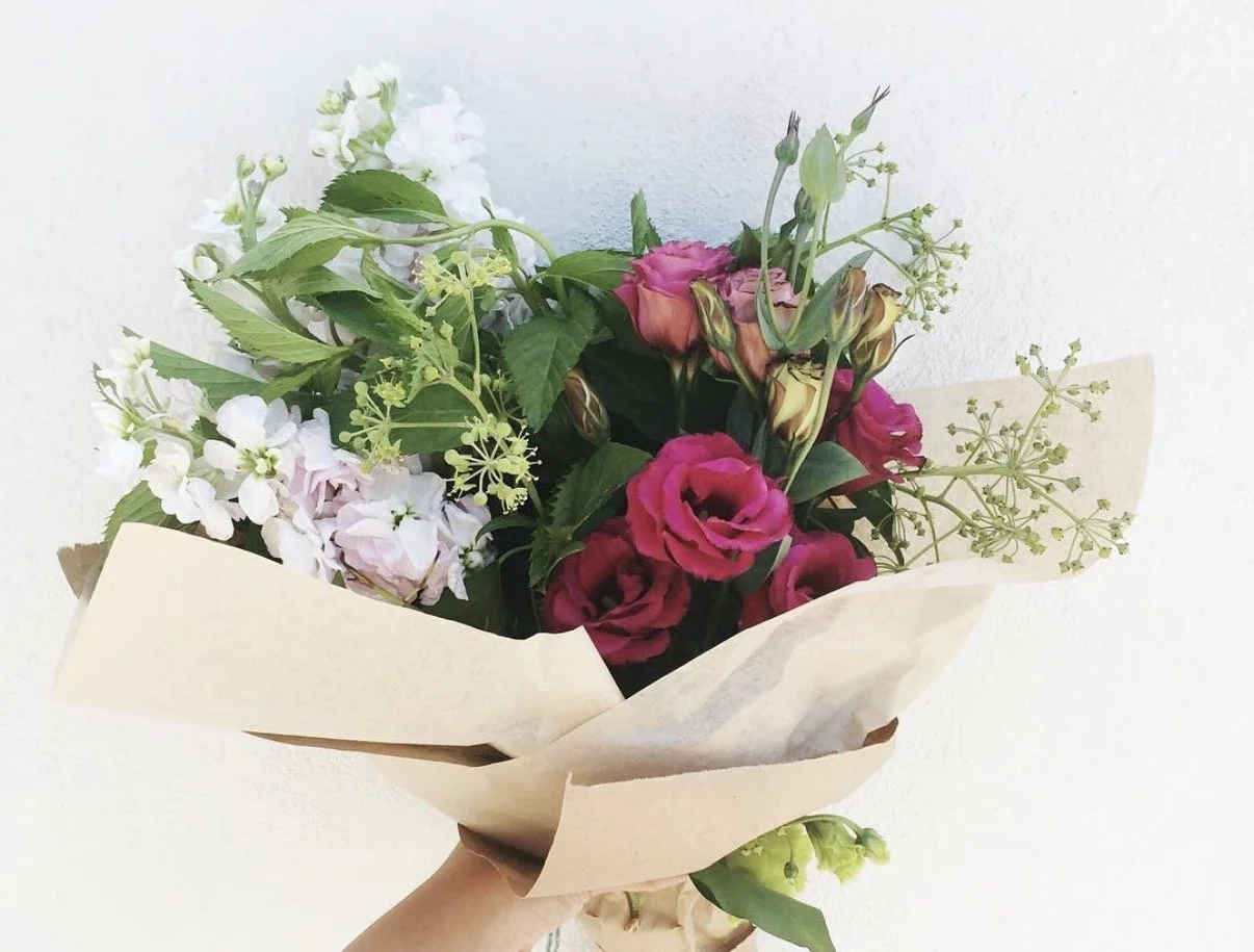 A hand holding a bouquet of pink, white, and green flowers wrapped in beige paper against a plain wall.