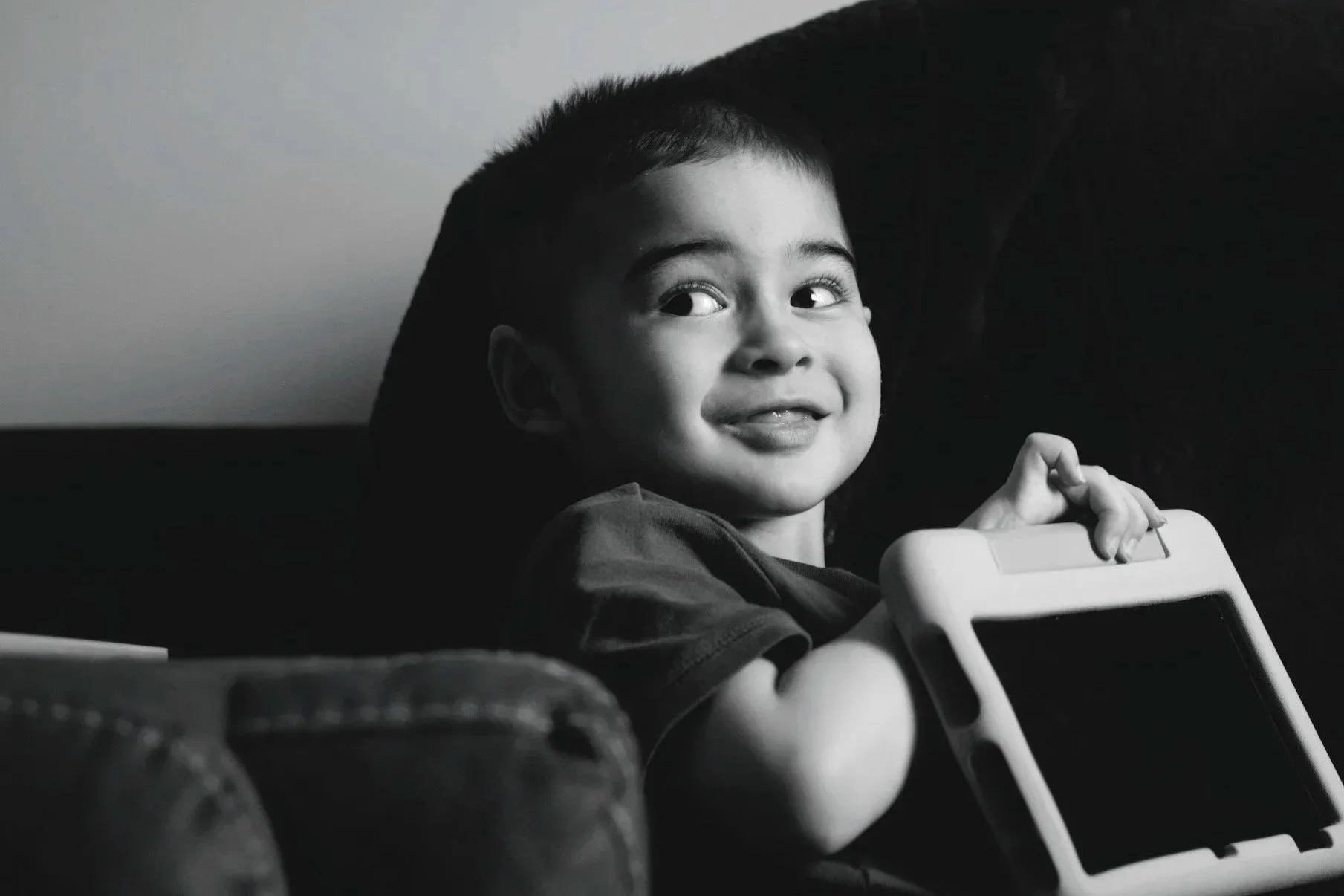 A young boy with short dark hair smiles and looks to the side, holding a tablet device while sitting on a dark background.