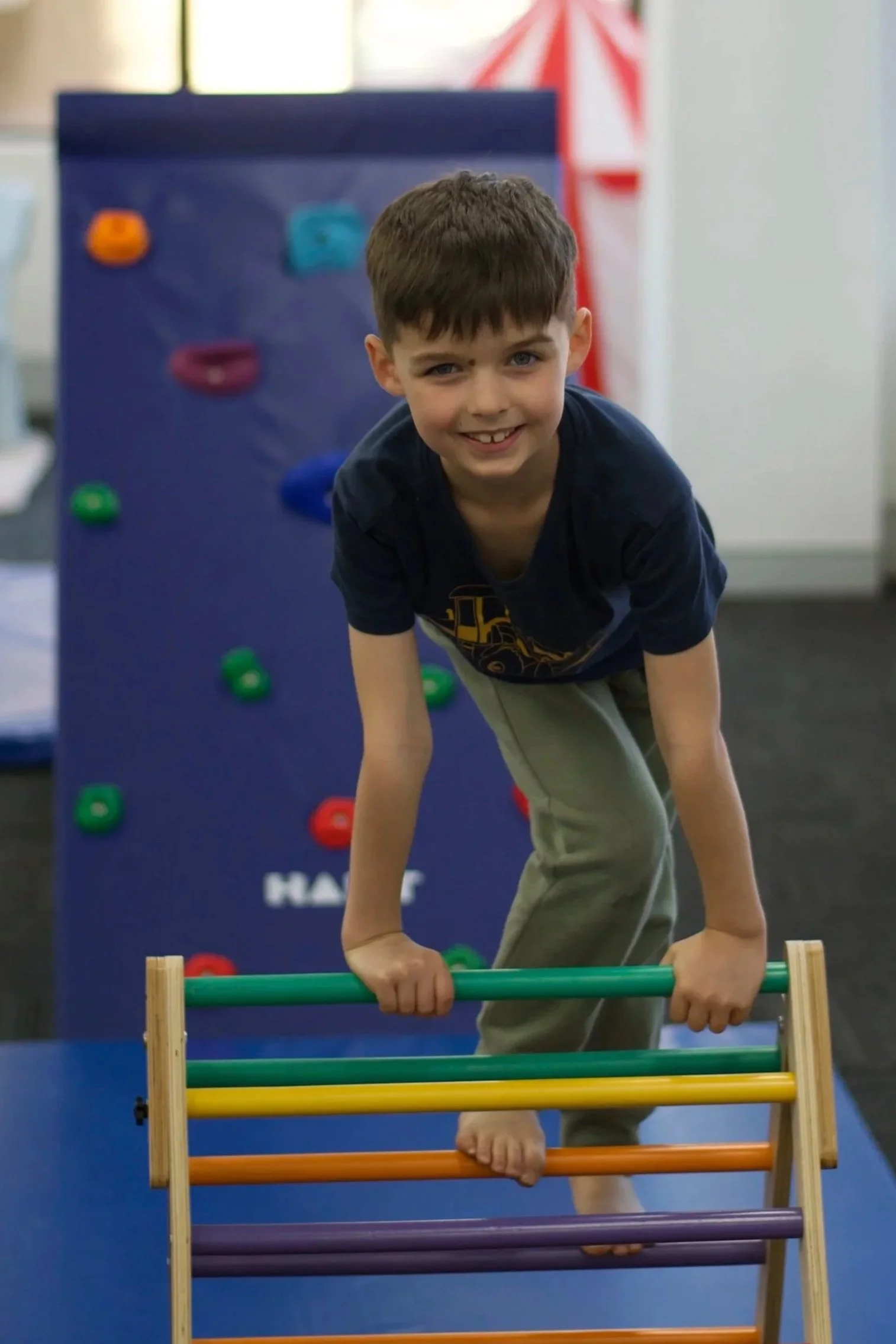 A young boy with brown hair smiling while crawling over a colorful Lego-like obstacle course at an indoor play area.